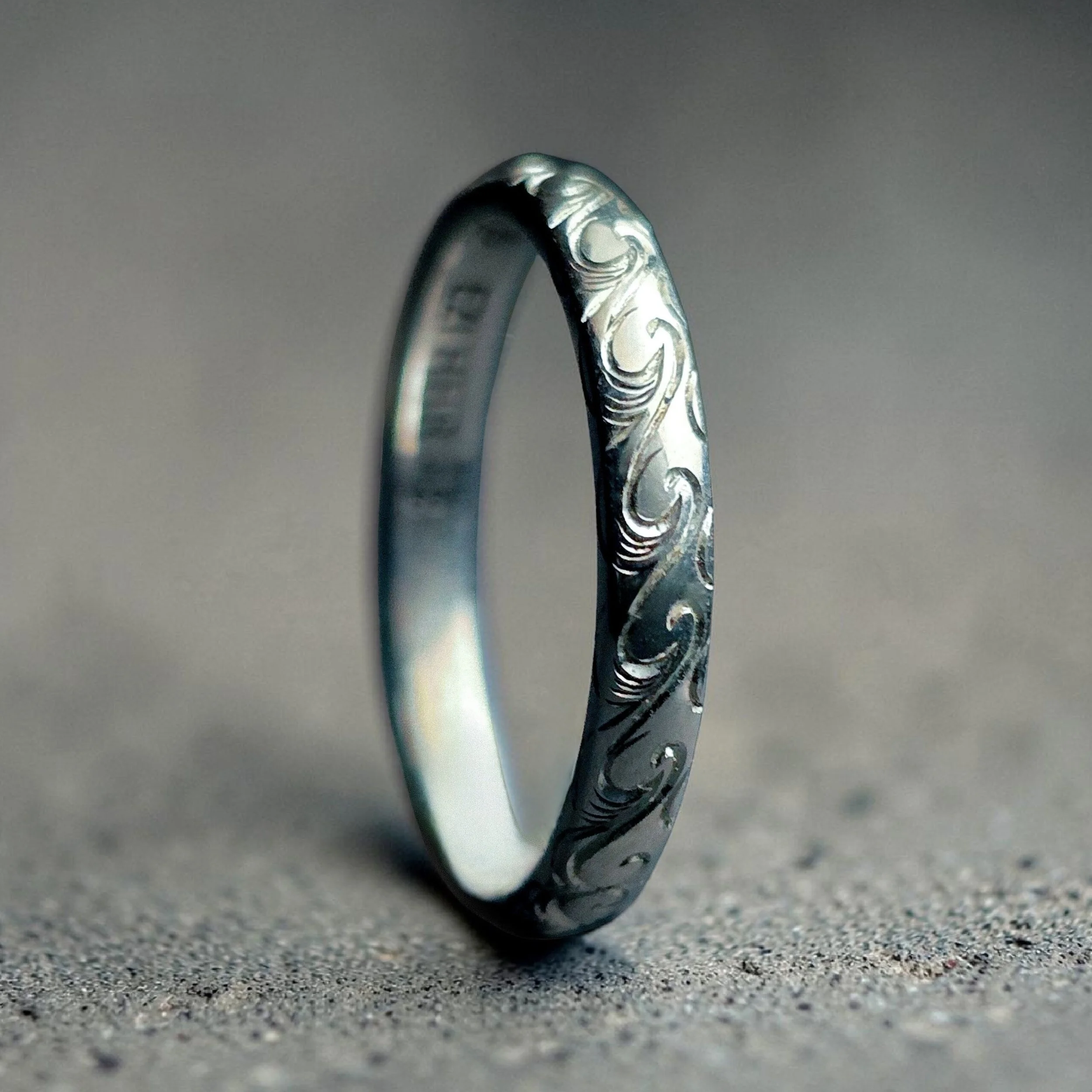 Close-up of a silver ring with intricate swirl patterns, lying on a textured surface.
