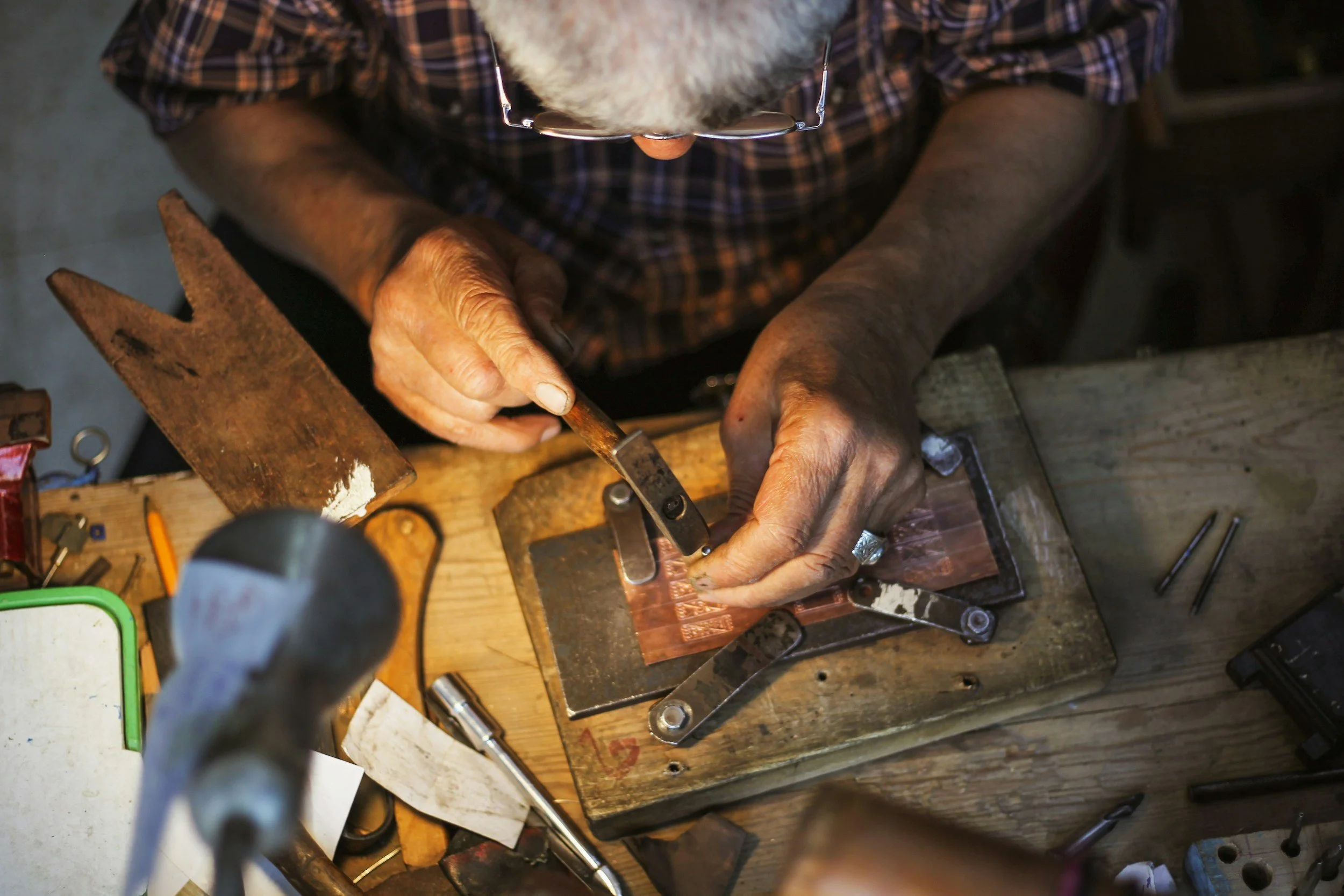 A person working at a woodworking or metalworking bench, closely focused on a project, with various tools and materials scattered around.