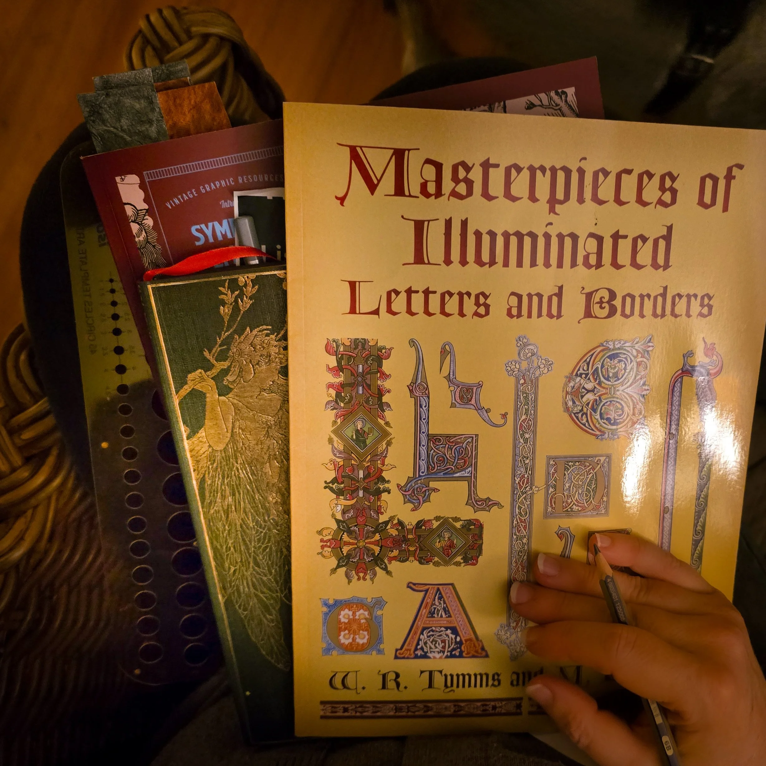 Person holding a book titled 'Masterpieces of Illuminated Letters and Borders' with other books and magazines nearby on a wooden surface.