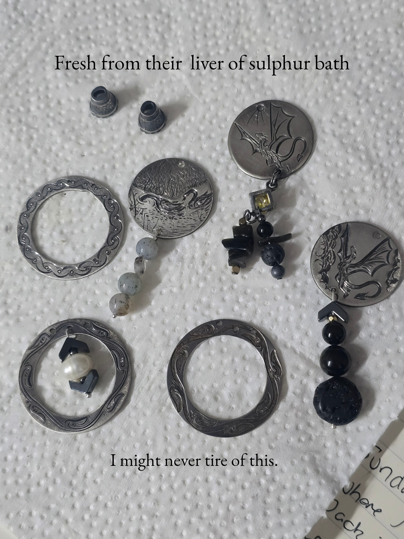 Close-up of various jewelry items on a white textured surface. The jewelry includes silver earrings, a necklace with a swan pendant, and other decorative pieces. Text overlays say, 'Fresh from their liver of sulphur bath' and 'I might never tire of this'.