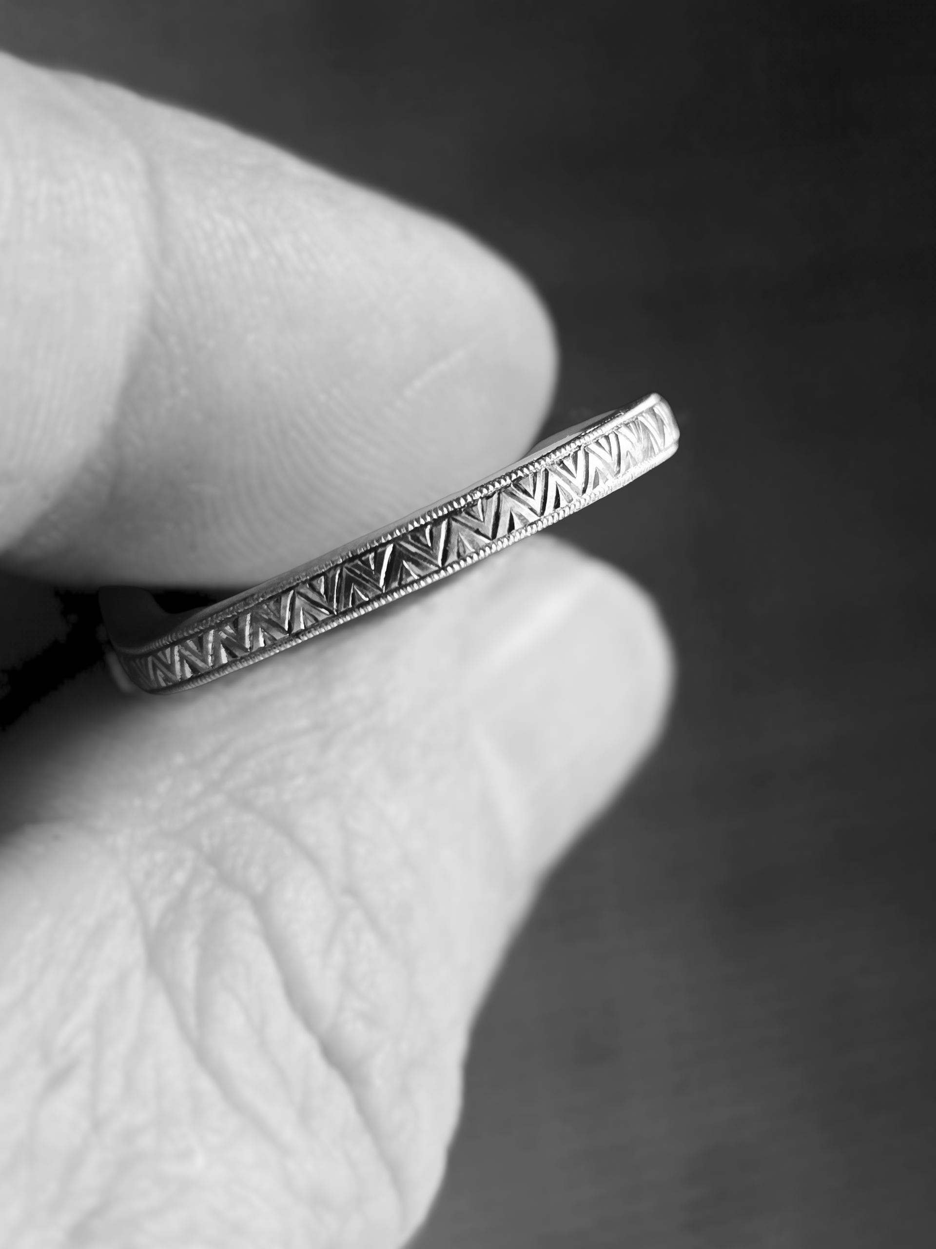 A person wearing a white glove holding a white gold ring with intricate geometric hand engravings, against a dark background.