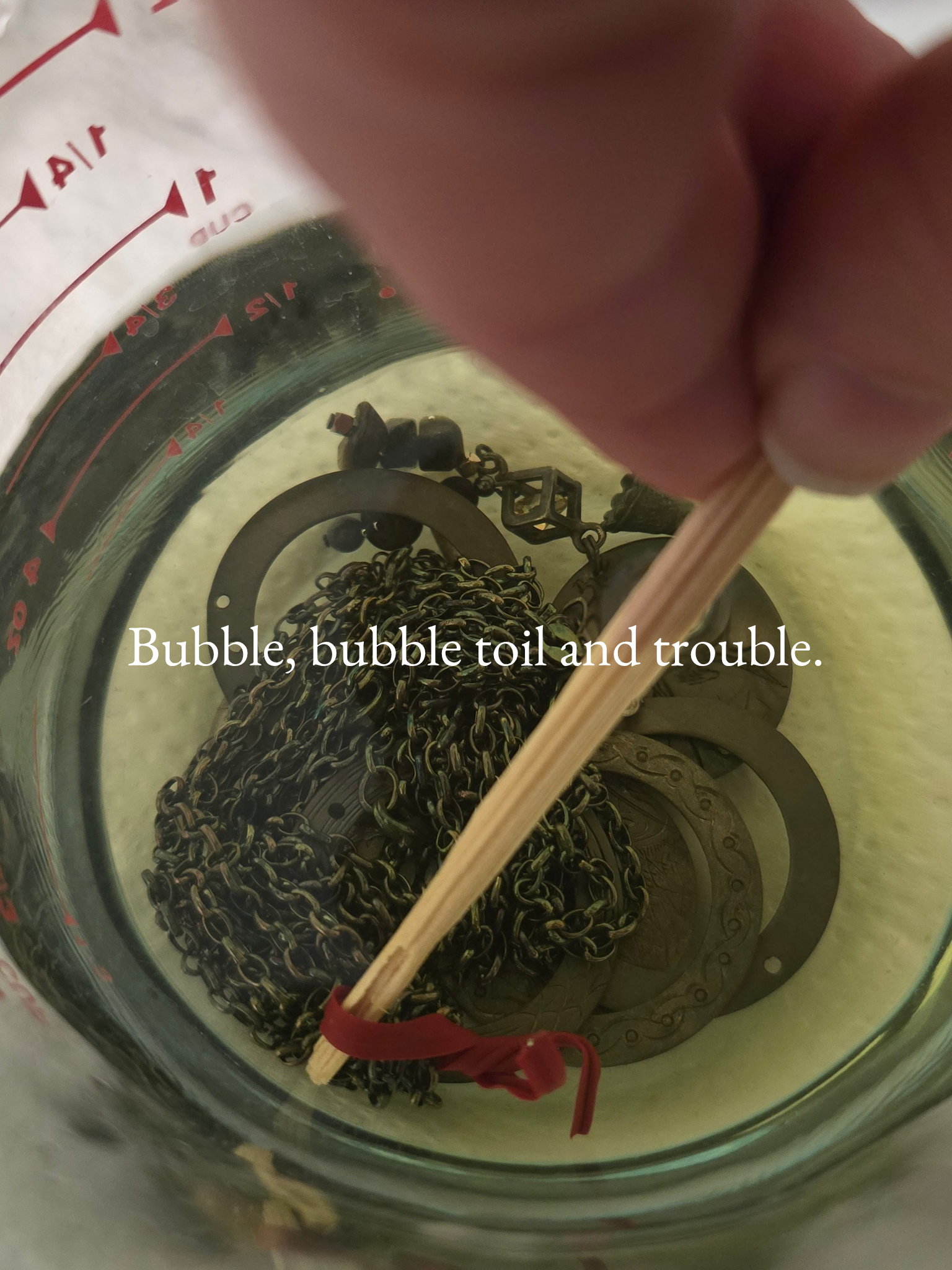 Inside a jar filled with black chain links, a red ribbon tied around a wooden stick, with other mechanical parts at the bottom. The text overlay says 'Bubble, bubble toil and trouble.'