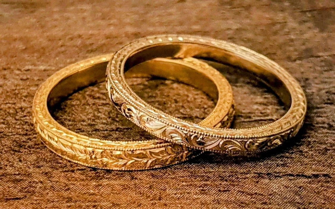 Two gold engagement or wedding rings with intricate hand engraved scroll and milgrain designs lying on a brown textured surface.