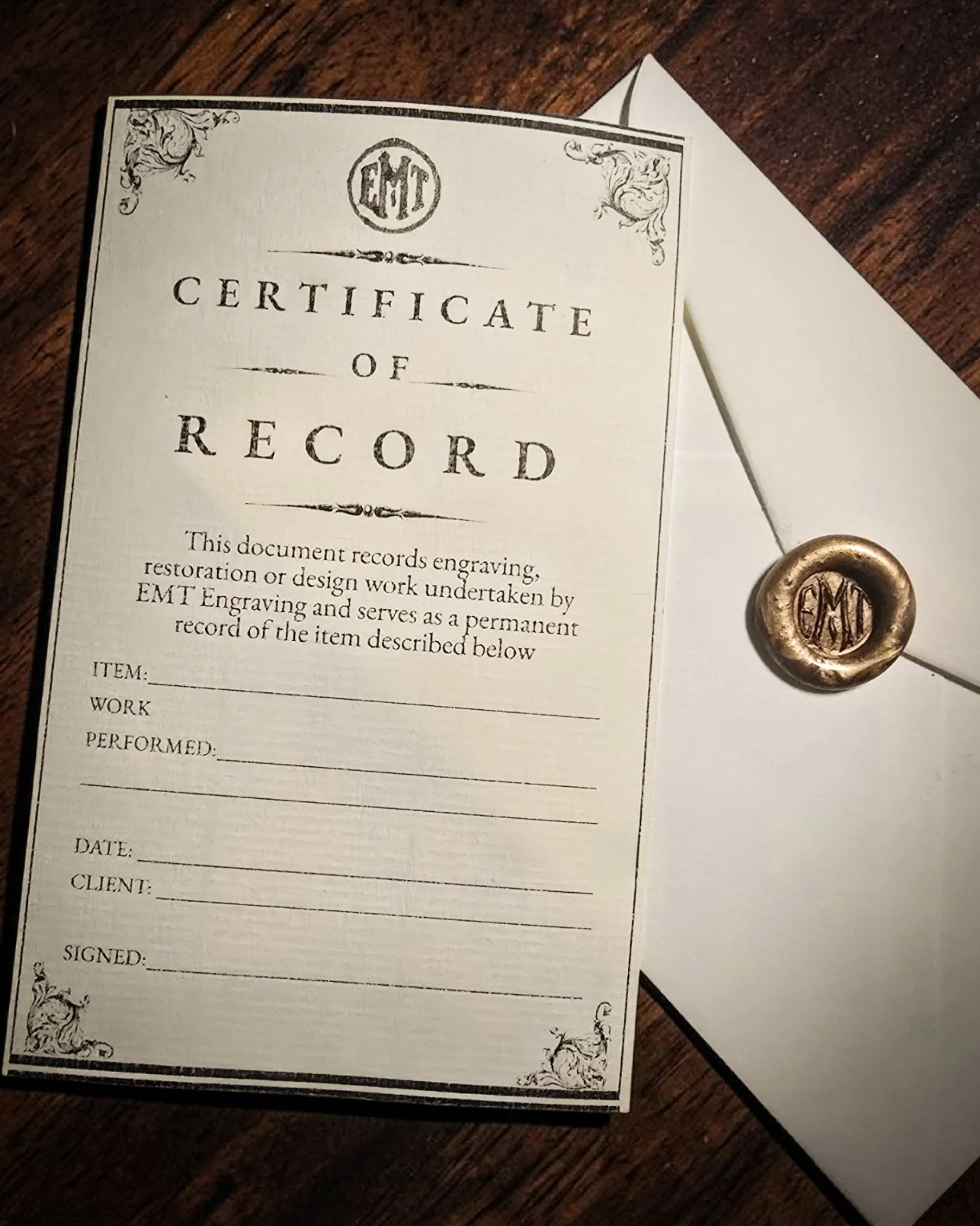 A vintage-style certificate of record with ornate decorative borders and a circular emblem at the top containing the initials 'EMT'. To the right of the certificate is a wax seal with the initials 'EMT'. The certificate appears to be resting on a wooden surface and features sections for item details, date, client, and signature.