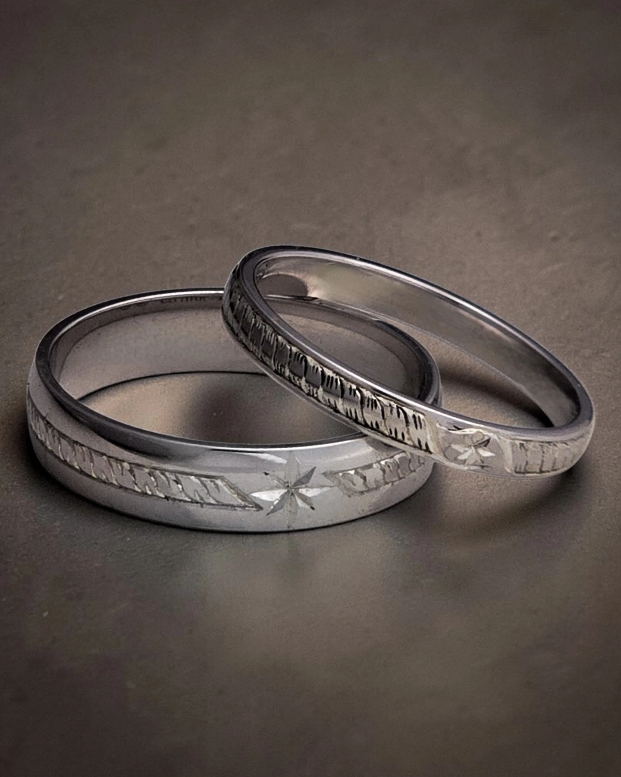 Close-up of intertwined silver wedding bands with custom, unique, bespoke hand engraved patterns, placed on a dark gray surface.