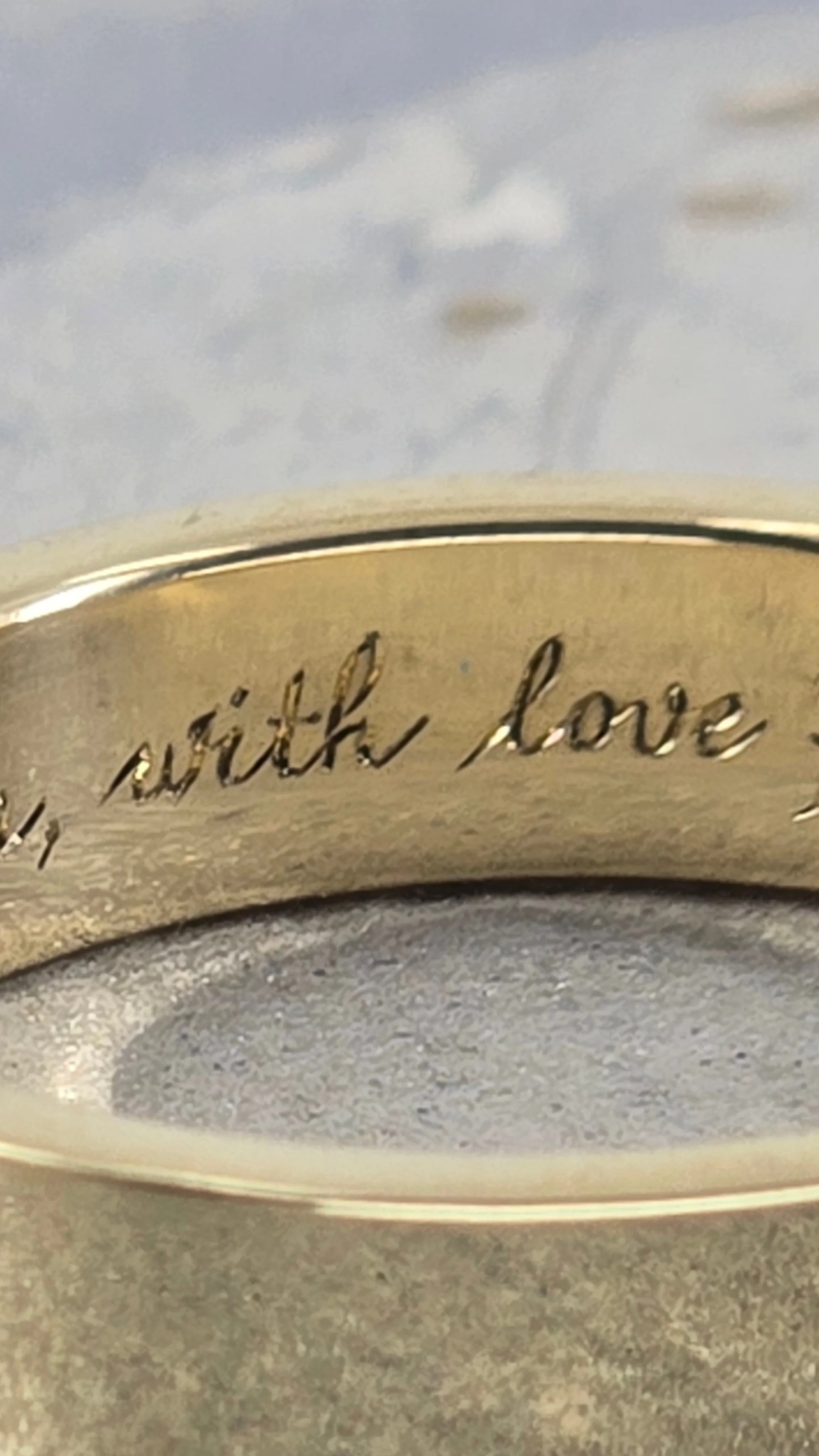 Close-up of a gold ring with the inscription "with love" hand engraved on it, placed on a textured gray surface with a cloudy background.