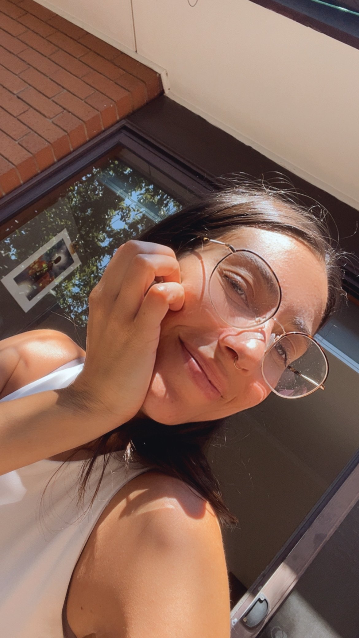 A woman with brown hair, wearing glasses, smiling and holding her glasses with one hand, standing outside near a brick building and a glass door, with trees reflected in the glass.