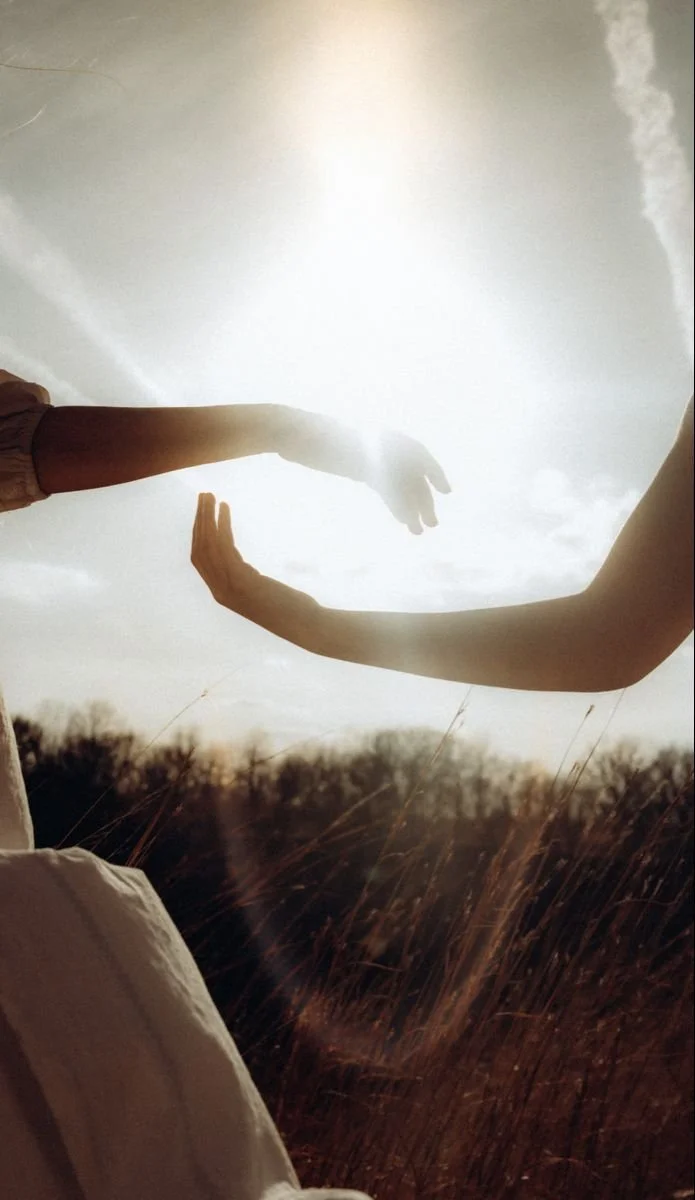 A person extending their arm and hand toward another person's arm, creating a visual illusion of the person in the background being touched by the finger of the person in the foreground, outdoors with grassy field and cloudy sky.