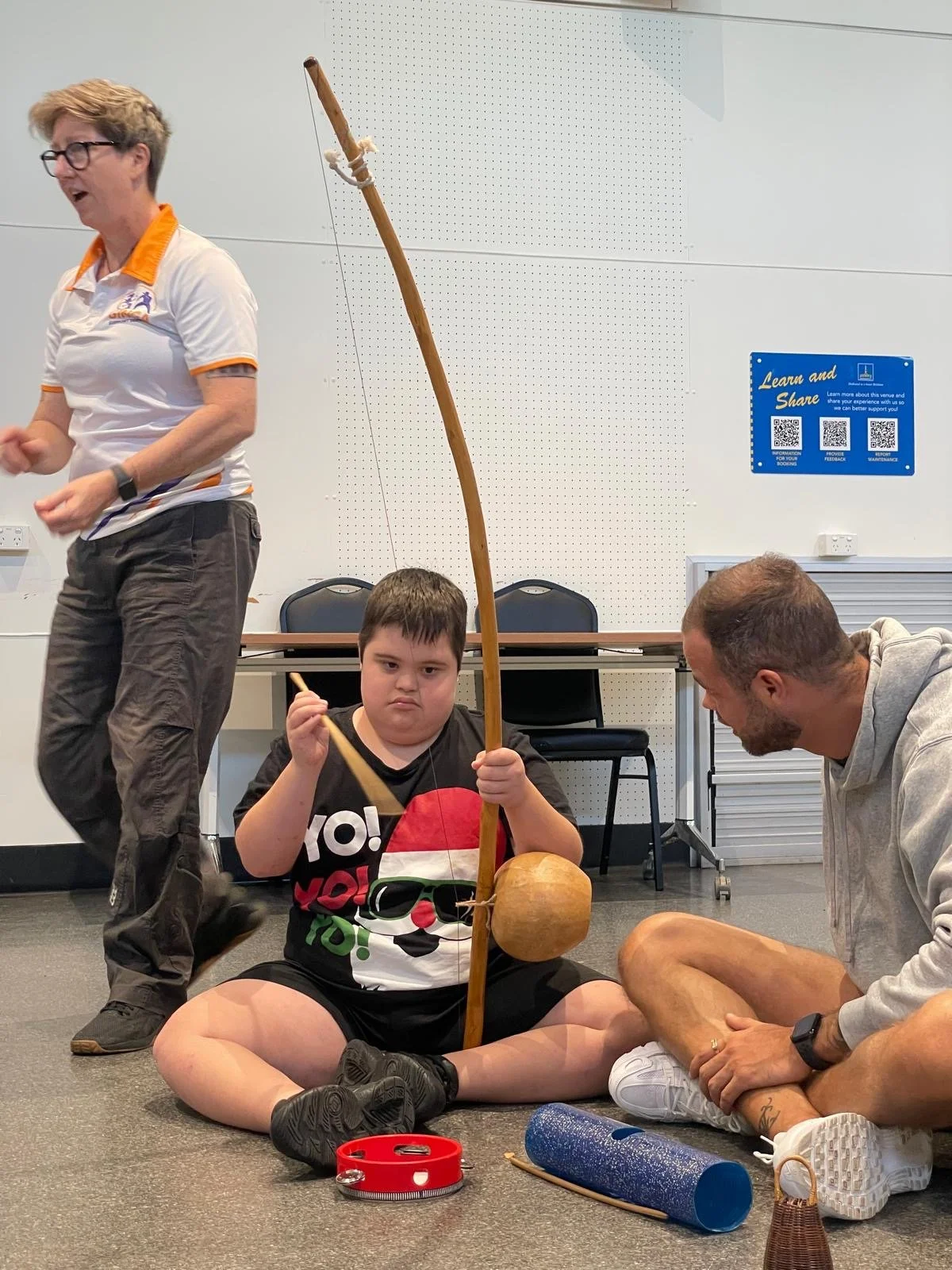 A disabled boy wearing a Christmas shirt and playing Brazilian instruments while sitting on the floor next to his support worker