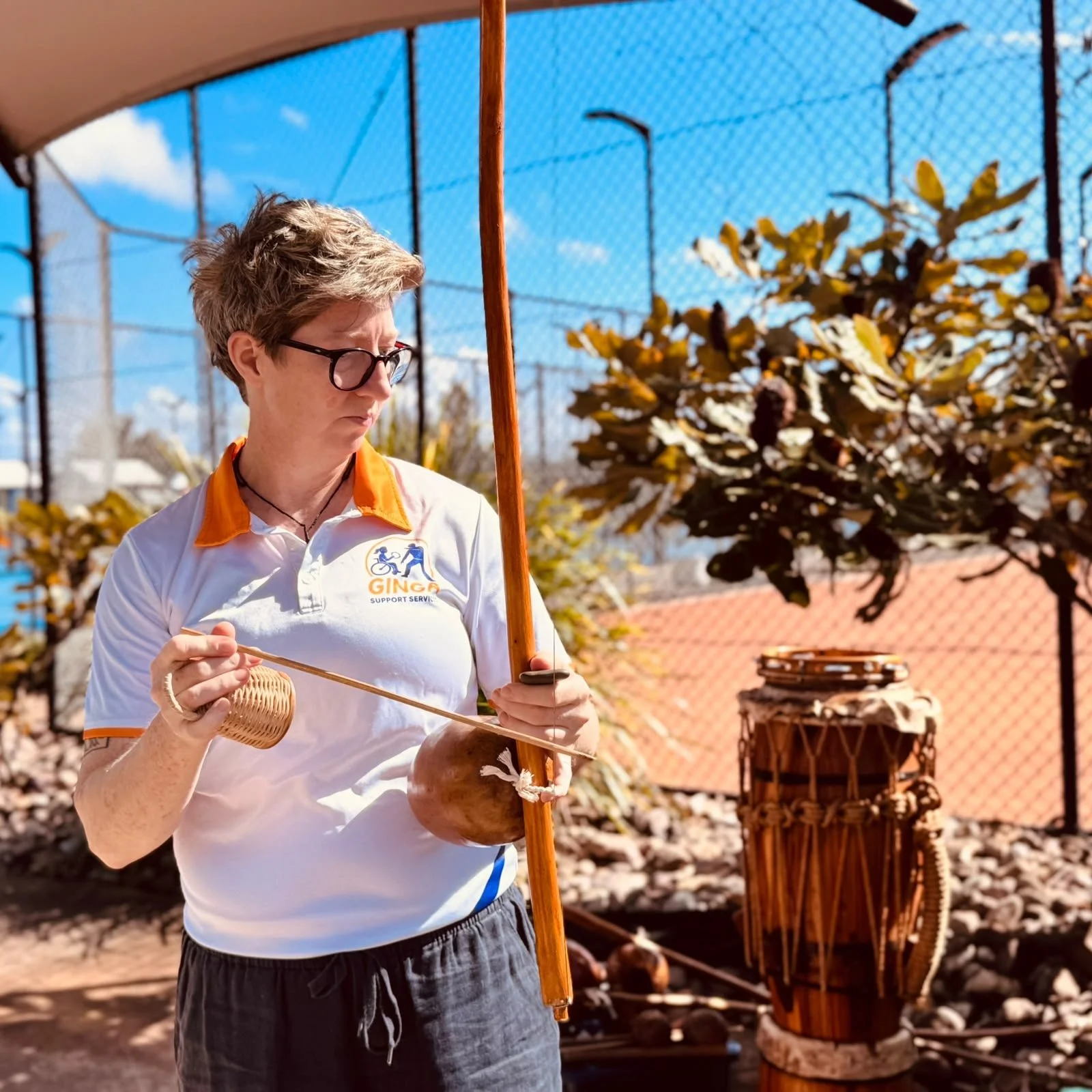 A woman wearing a white and orange trim polo shirt. She is holding Brazilian instruments used in Capoeira and is heavily focused on the task she is doing