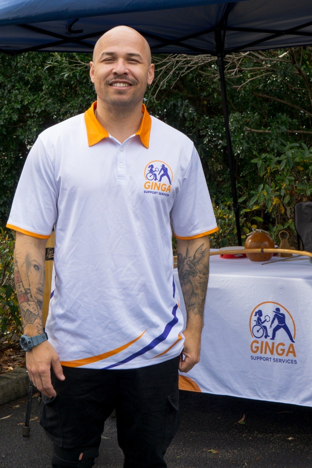 A bald man with a white and orange trim polo shirt. He has tattoos on his arms and a moustache.