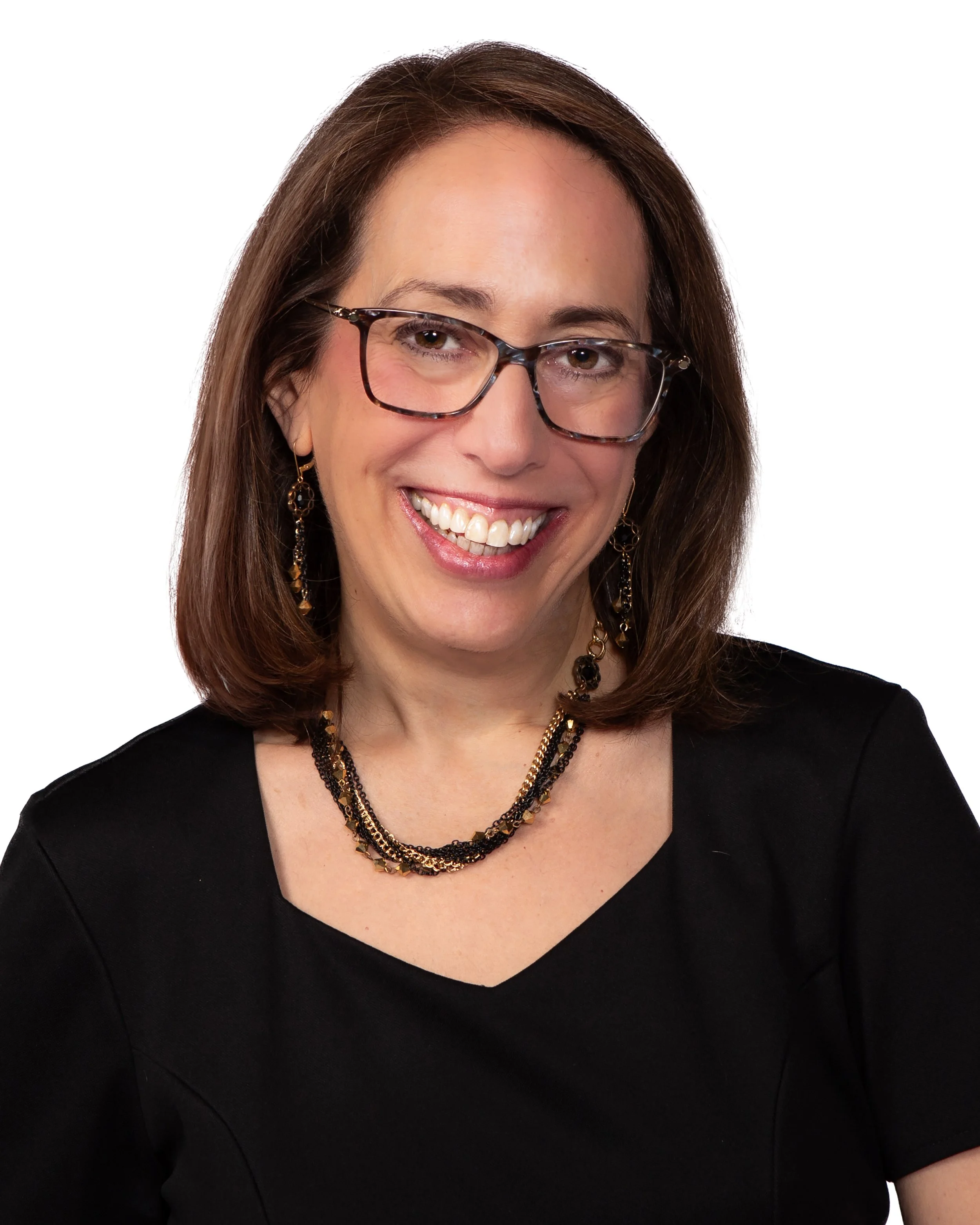 A woman with brown hair wearing glasses, earrings, and a layered necklace, dressed in a black top, smiling against a white background.