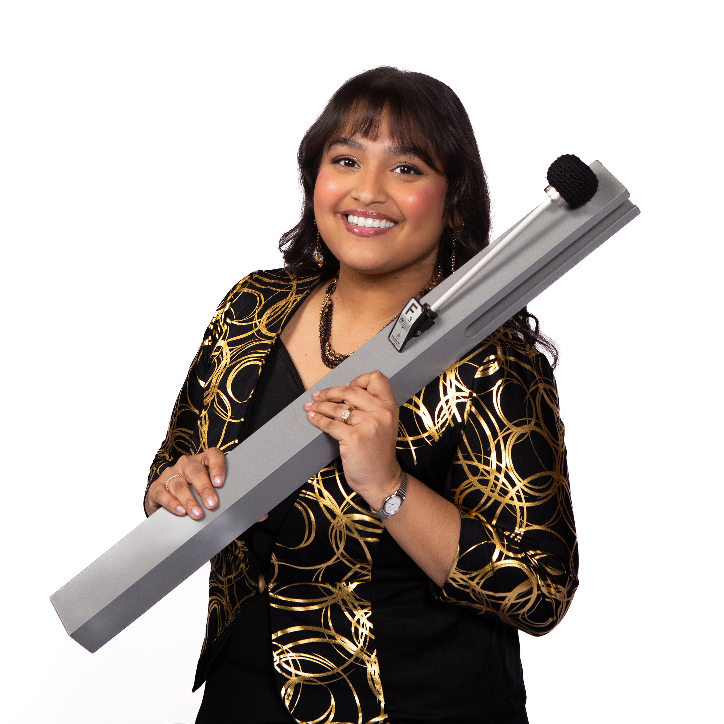A woman with dark brown hair, smiling, holding a large chime, wearing a black outfit with a gold geometric pattern.