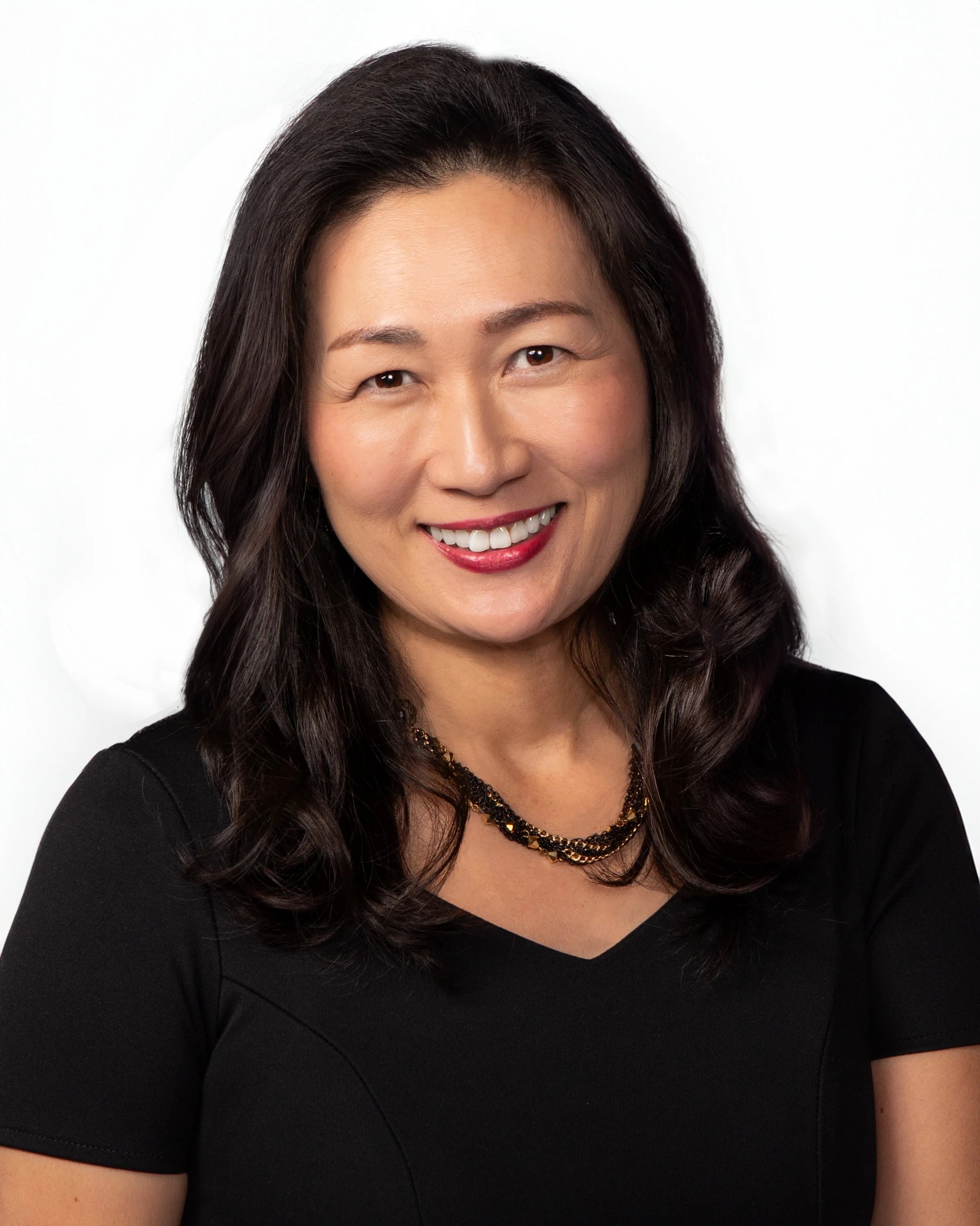 Close-up of a woman with long dark hair, smiling, wearing a black top and a layered gold necklace, against a plain white background.