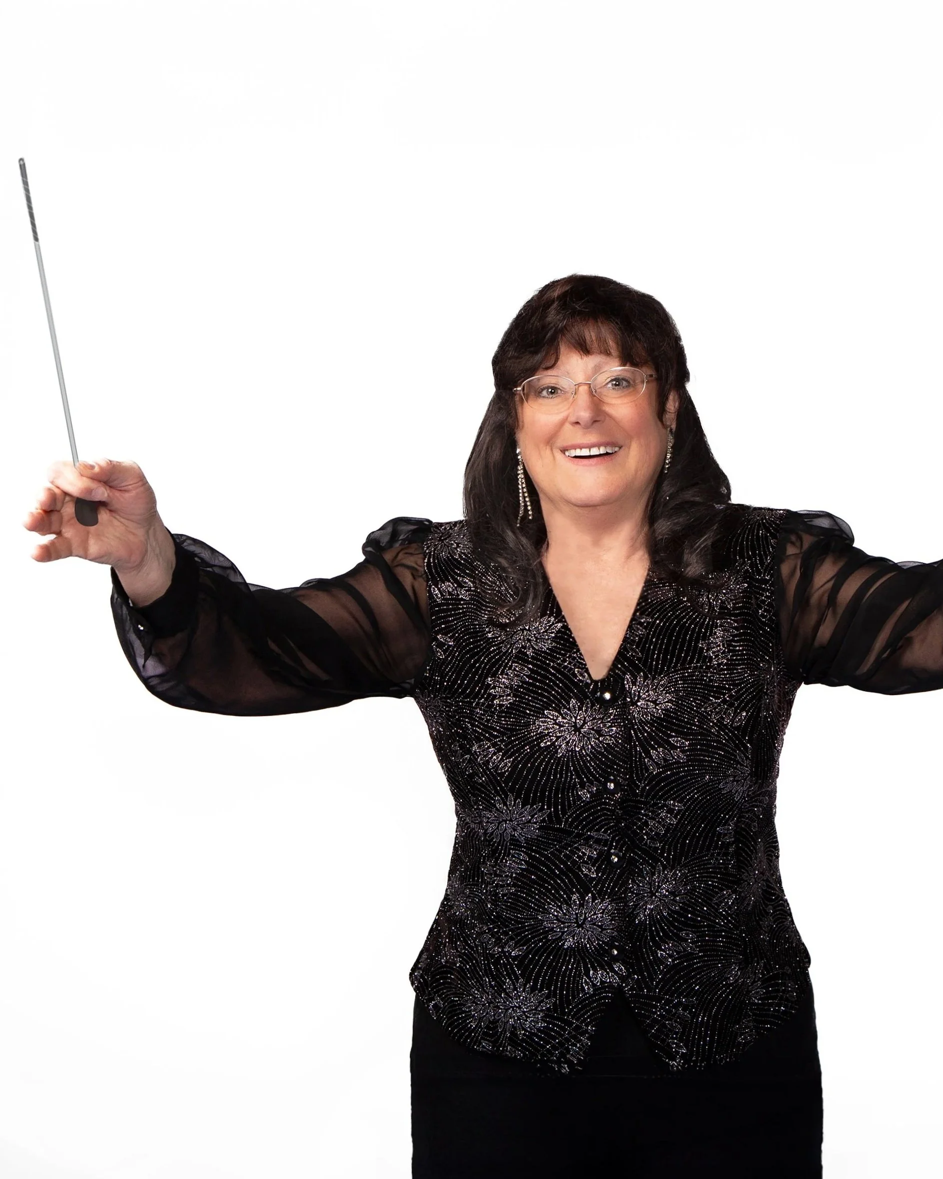 A woman with dark hair and glasses, wearing a black dress with shiny, floral embroidery, smiling and holding a thin, black and silver baton.