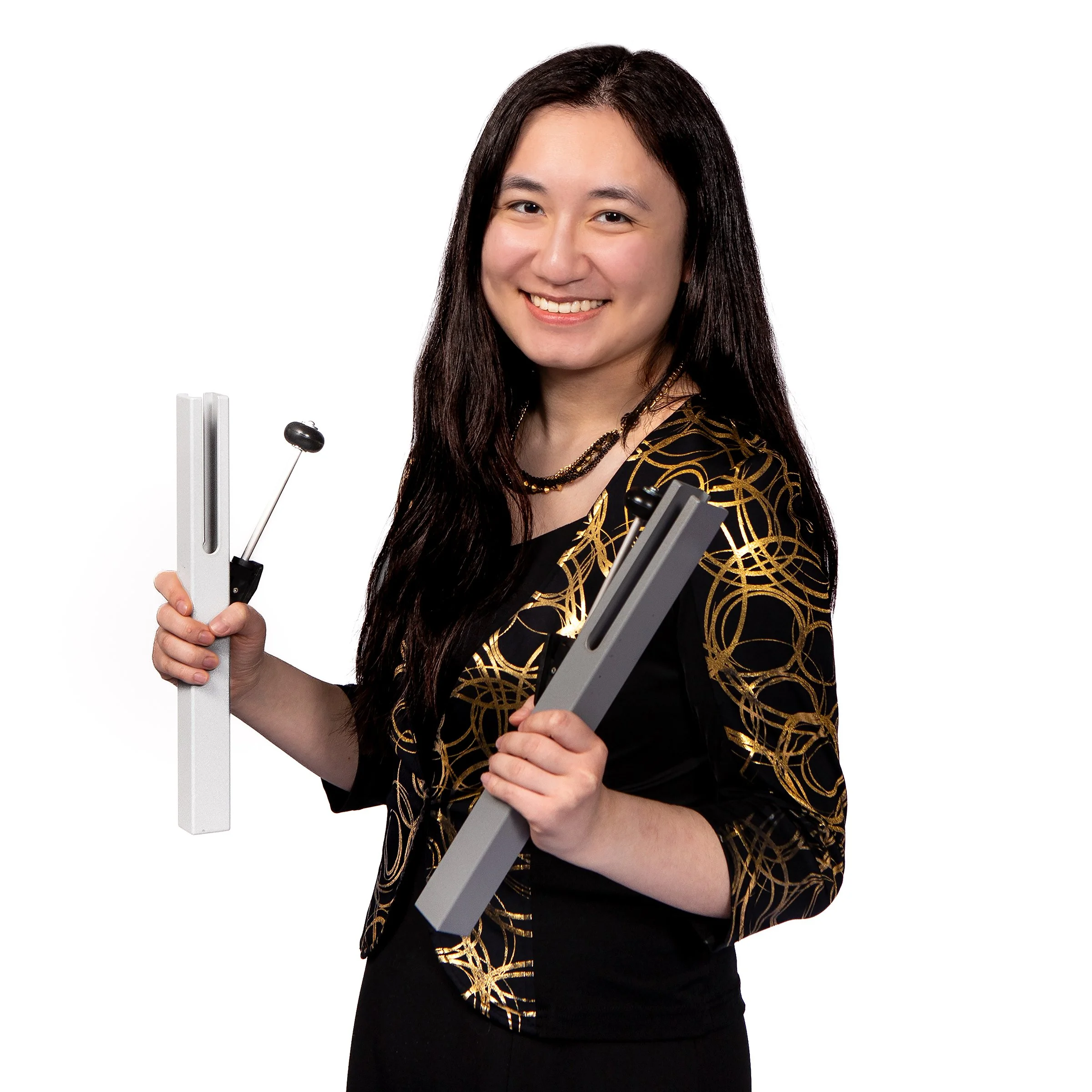 A woman with long dark hair smiling, wearing a black dress with gold patterns, holding various communication or electronic devices against a plain white background.
