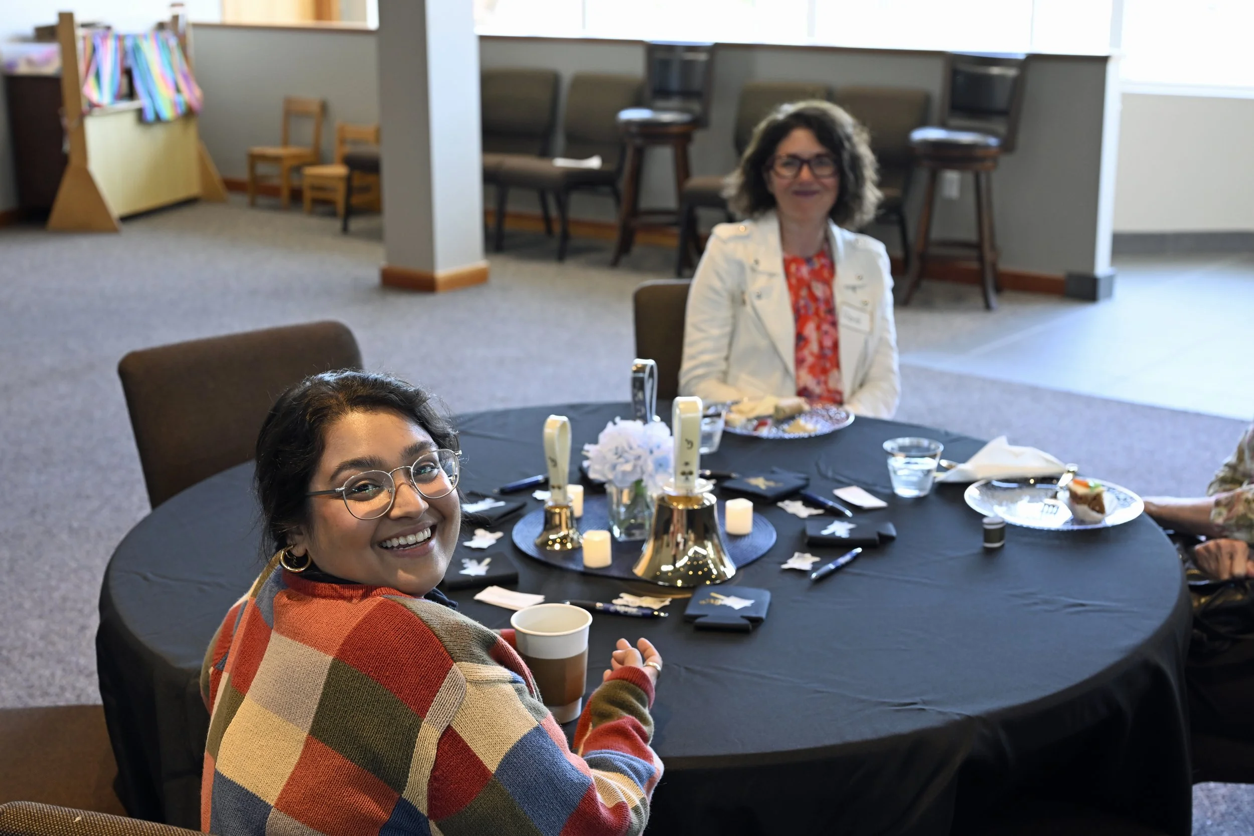 Smiling woman with glasses and a colorful checkered sweater sitting at a table with food and drinks, in a room with chairs and windows in the background.