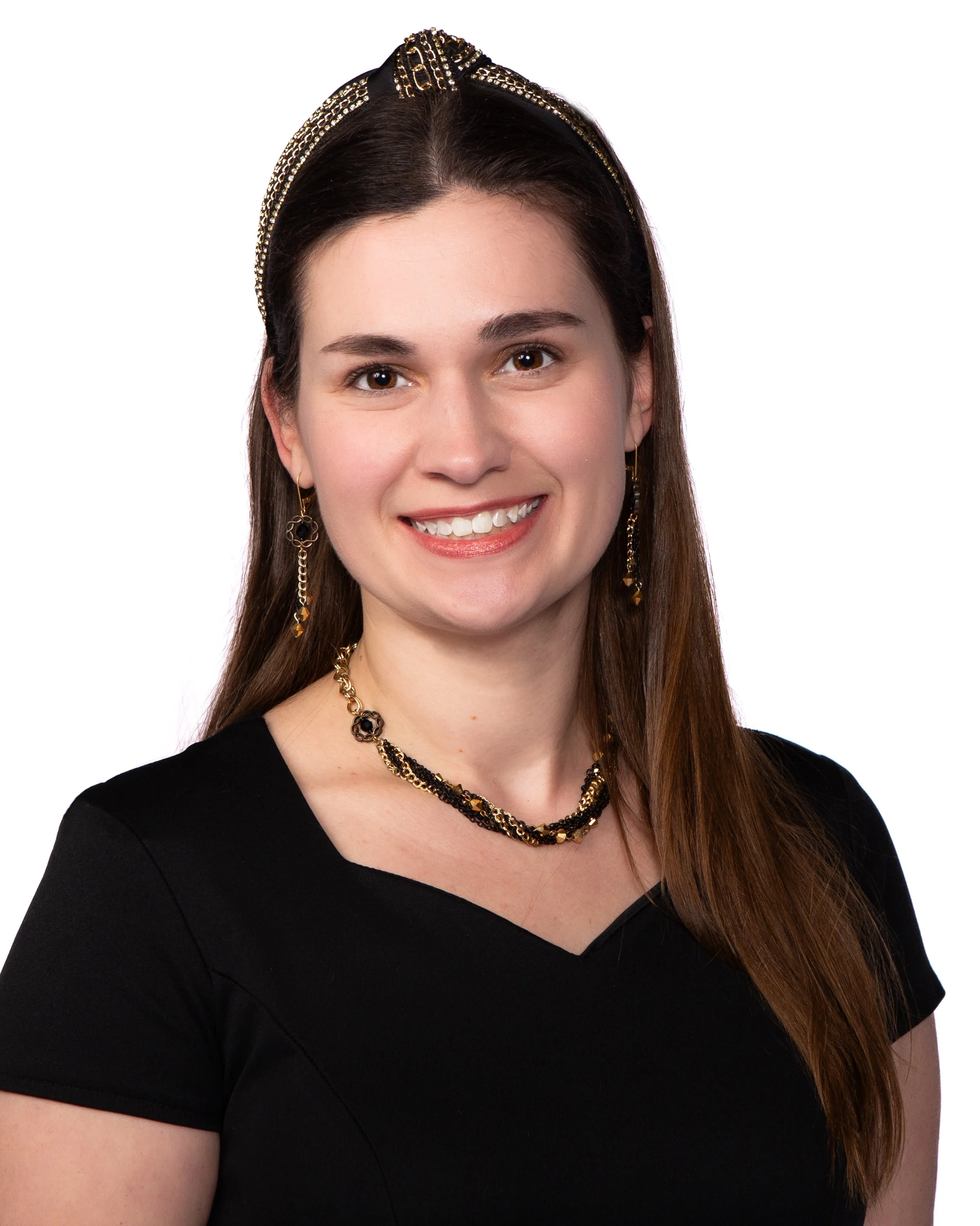 Close-up of a smiling woman with long brown hair, wearing a black top, gold and black jewelry, and a black headband with gold accents, against a white background.