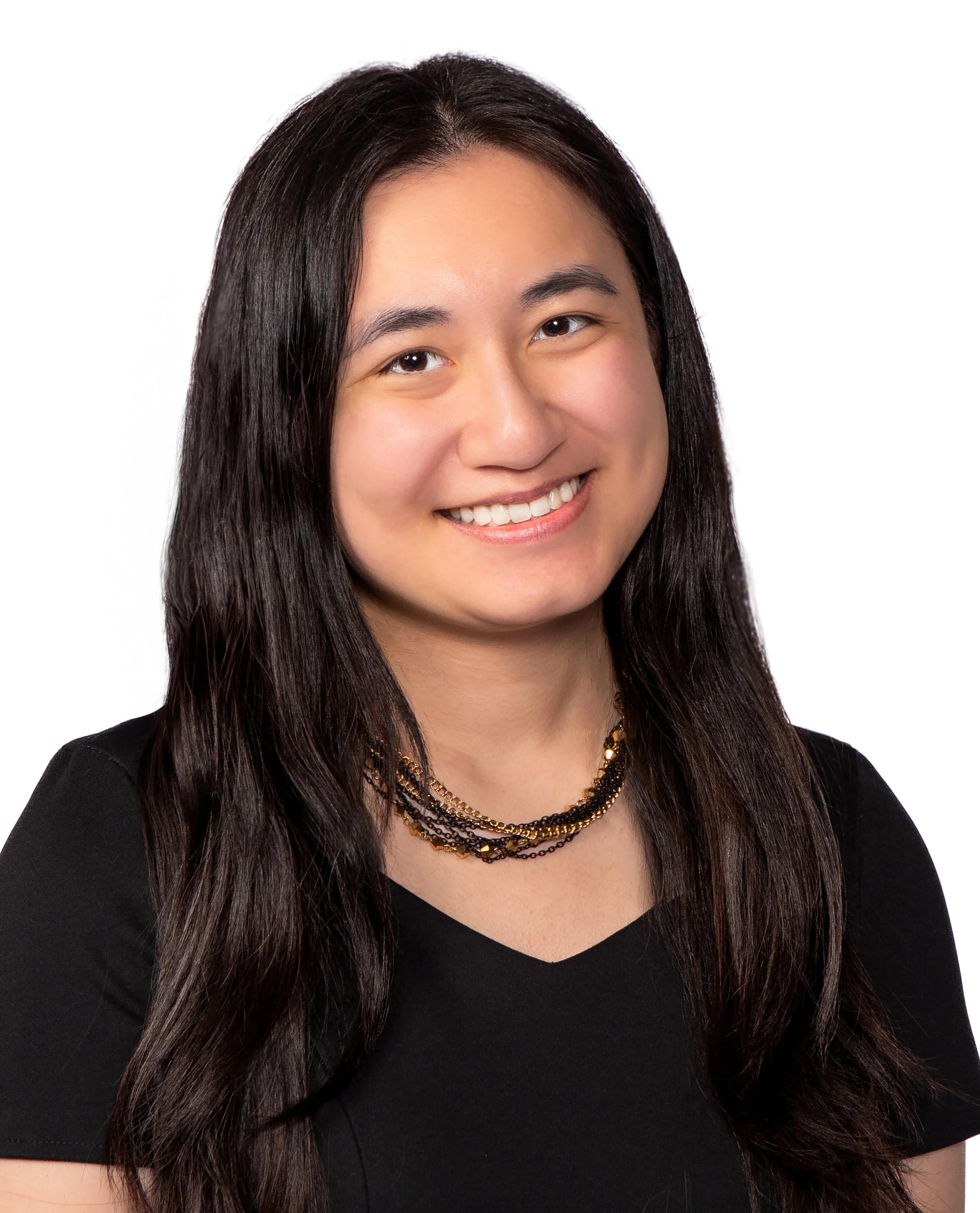 Close-up of a woman with long dark hair smiling, wearing a black V-neck top and layered gold and black necklaces against a white background.