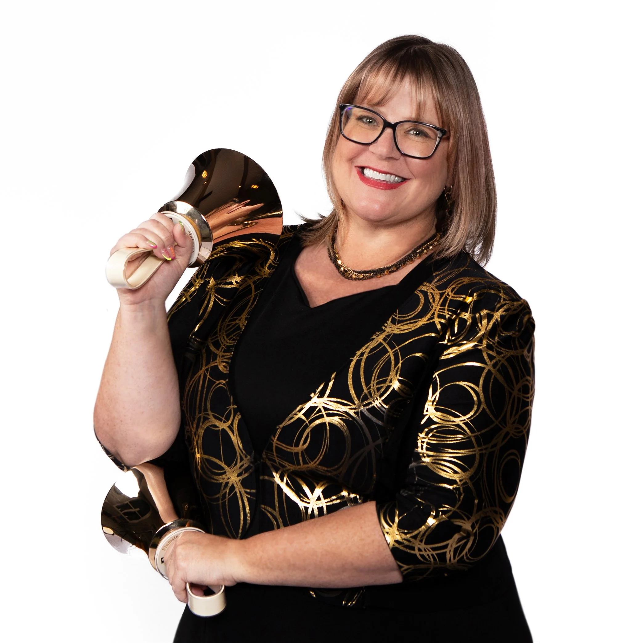 A woman with shoulder-length brown hair, wearing glasses, smiling, and holdinghandbells, standing against a white background, wearing a black outfit with gold patterns and jewelry.