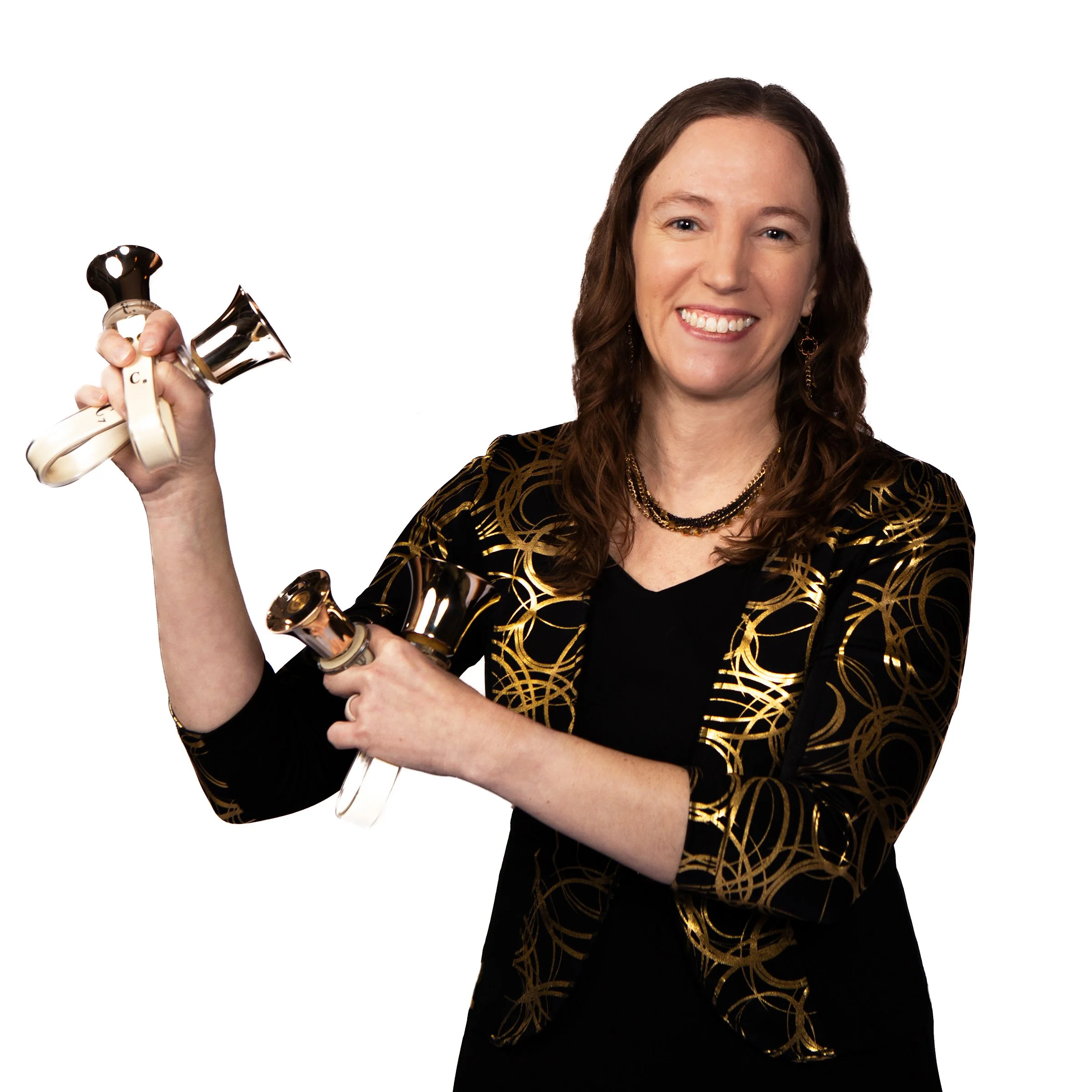 A woman smiling and holding handbells in each hand, wearing a black and gold patterned top, gold jewelry, with brown hair and a white background.