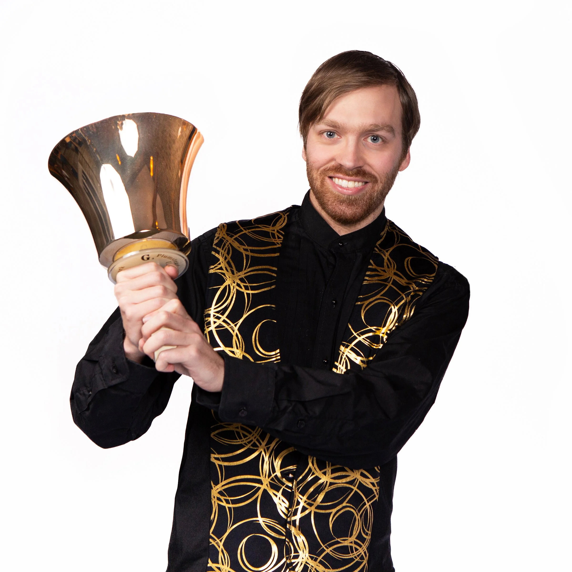 A man with short brown hair and a beard, smiling, holding a large handbell, wearing a black shirt with a gold swirling pattern, against a plain white background.