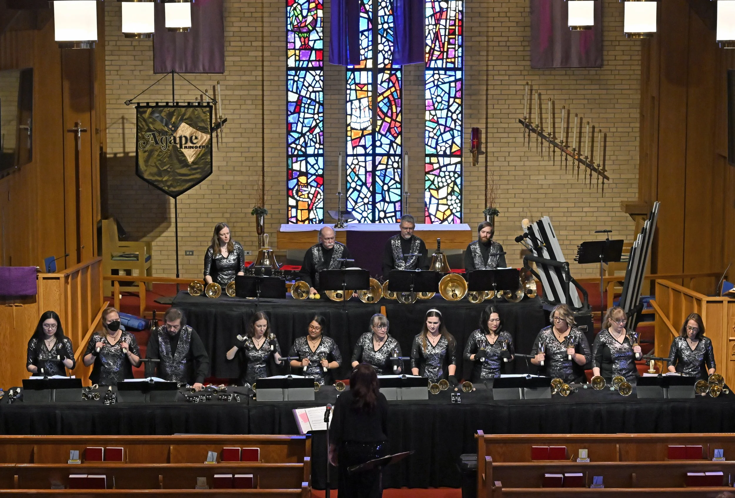 A handbell choir performing in a church with stained glass windows and wooden decor. The choir members are dressed in black with silver patterns, and are holding or standing near handbells. There is a conductor in front of them.