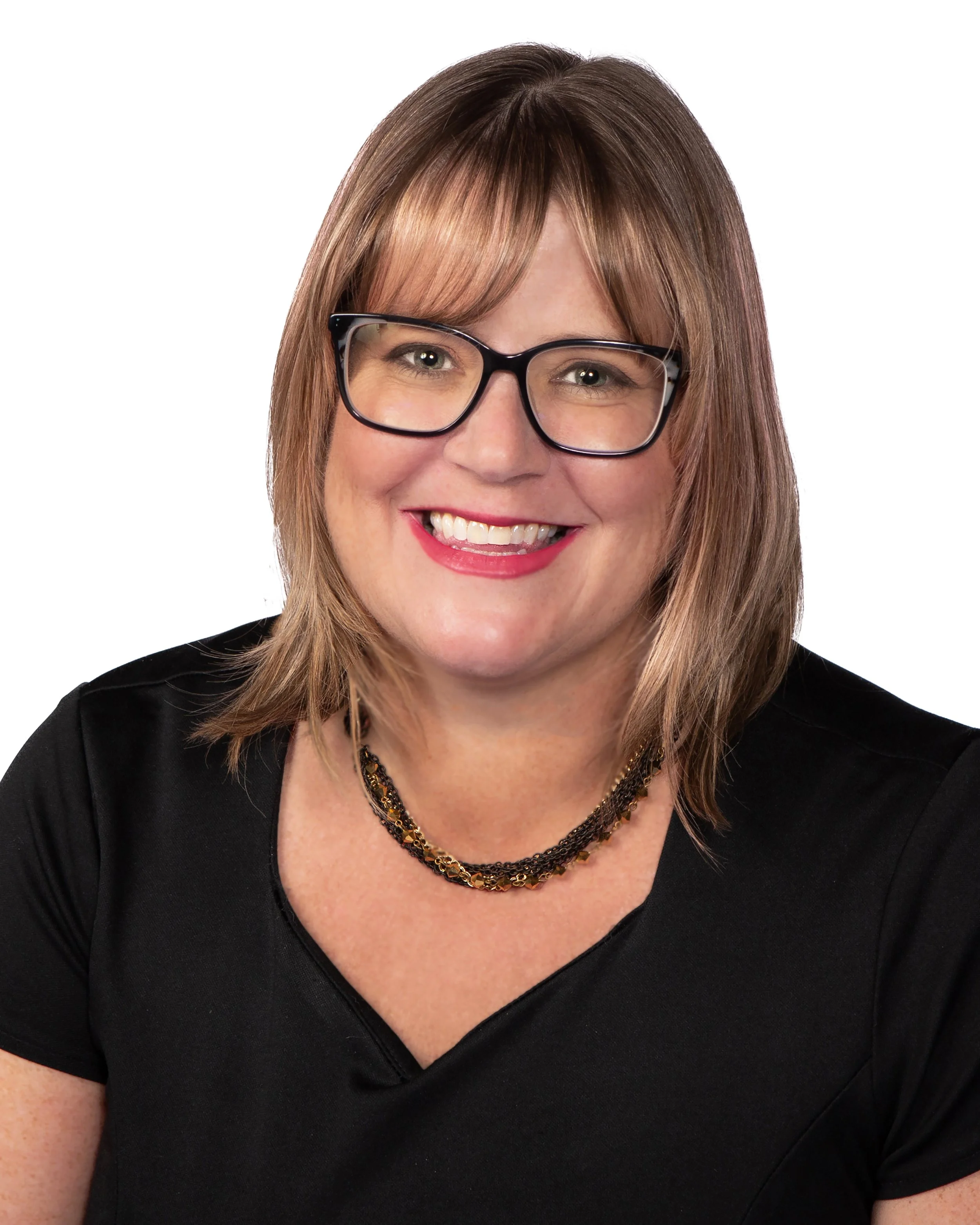 Close-up of a woman with brown hair, black-rimmed glasses, smiling, wearing a black top and layered necklaces, against a white background.