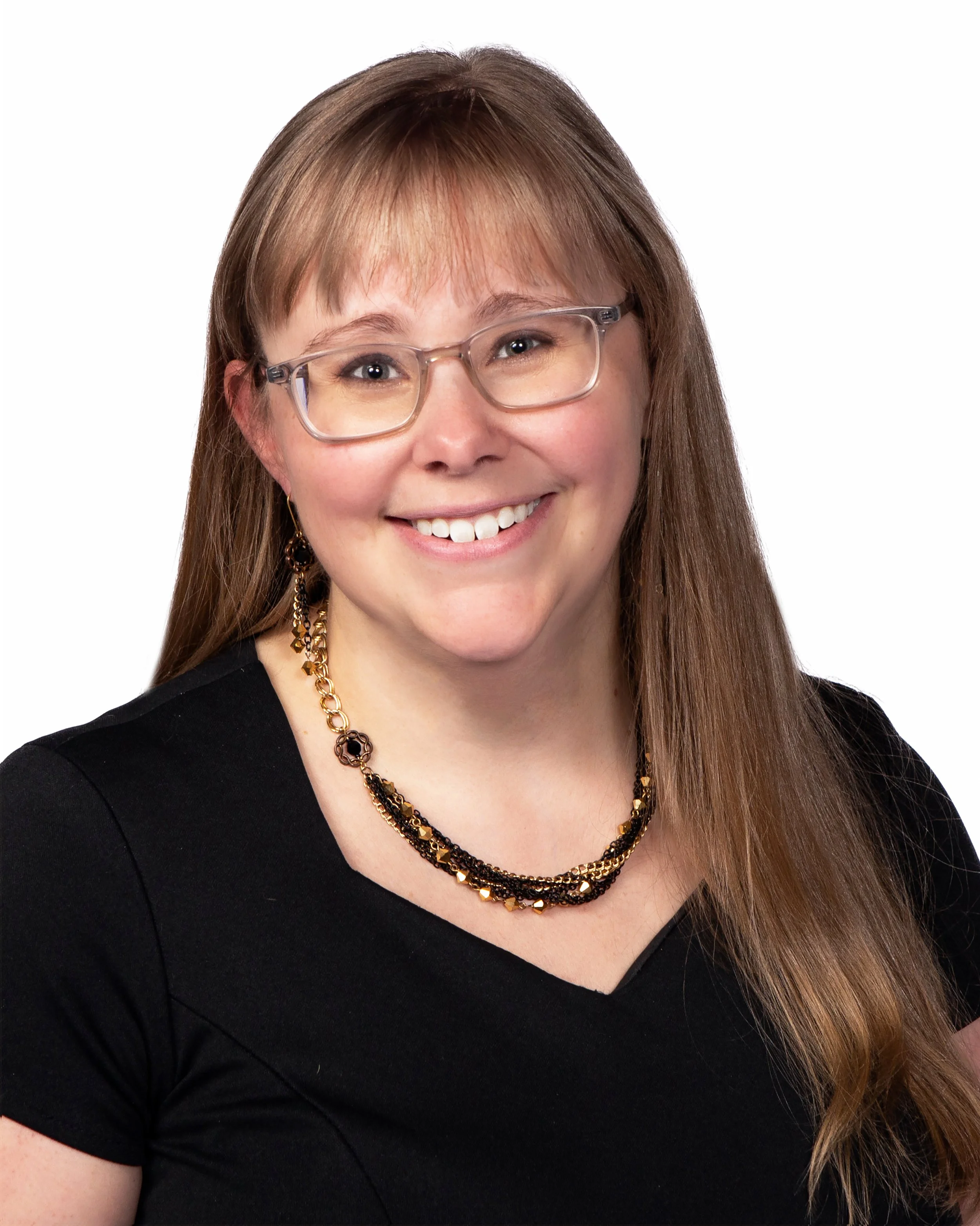 A woman with long brown hair, wearing glasses, a black top, and layered gold and black necklaces, smiling against a plain white background.