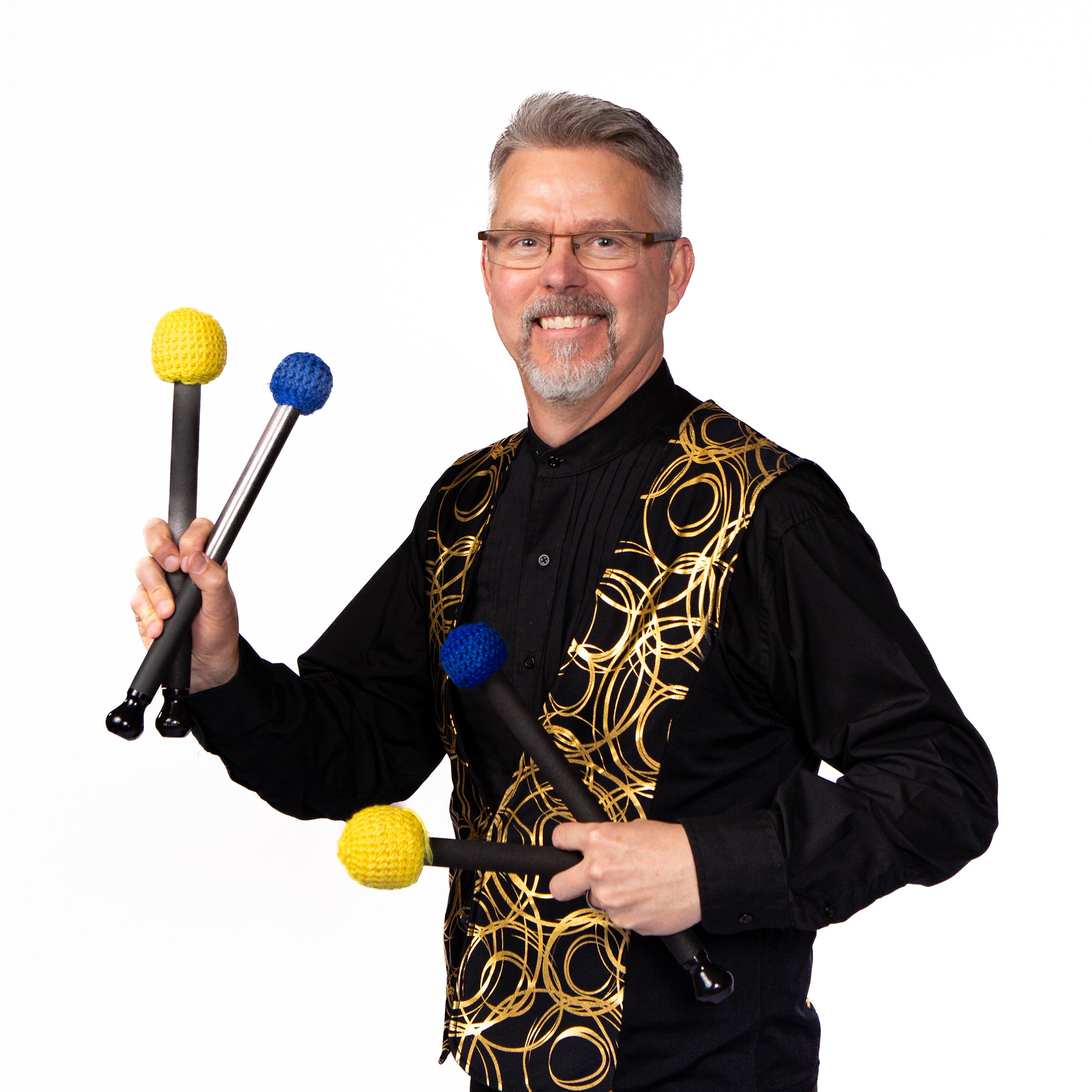 A smiling man with gray hair and glasses, wearing a black shirt and a black jacket with gold swirl patterns, holding three mallets with yellow and blue heads against a white background.