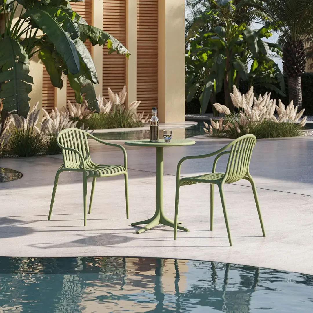 Two green slatted chairs and a round café table sit beside a reflective pool, surrounded by tropical plants and warm modern architecture, creating a serene mood.