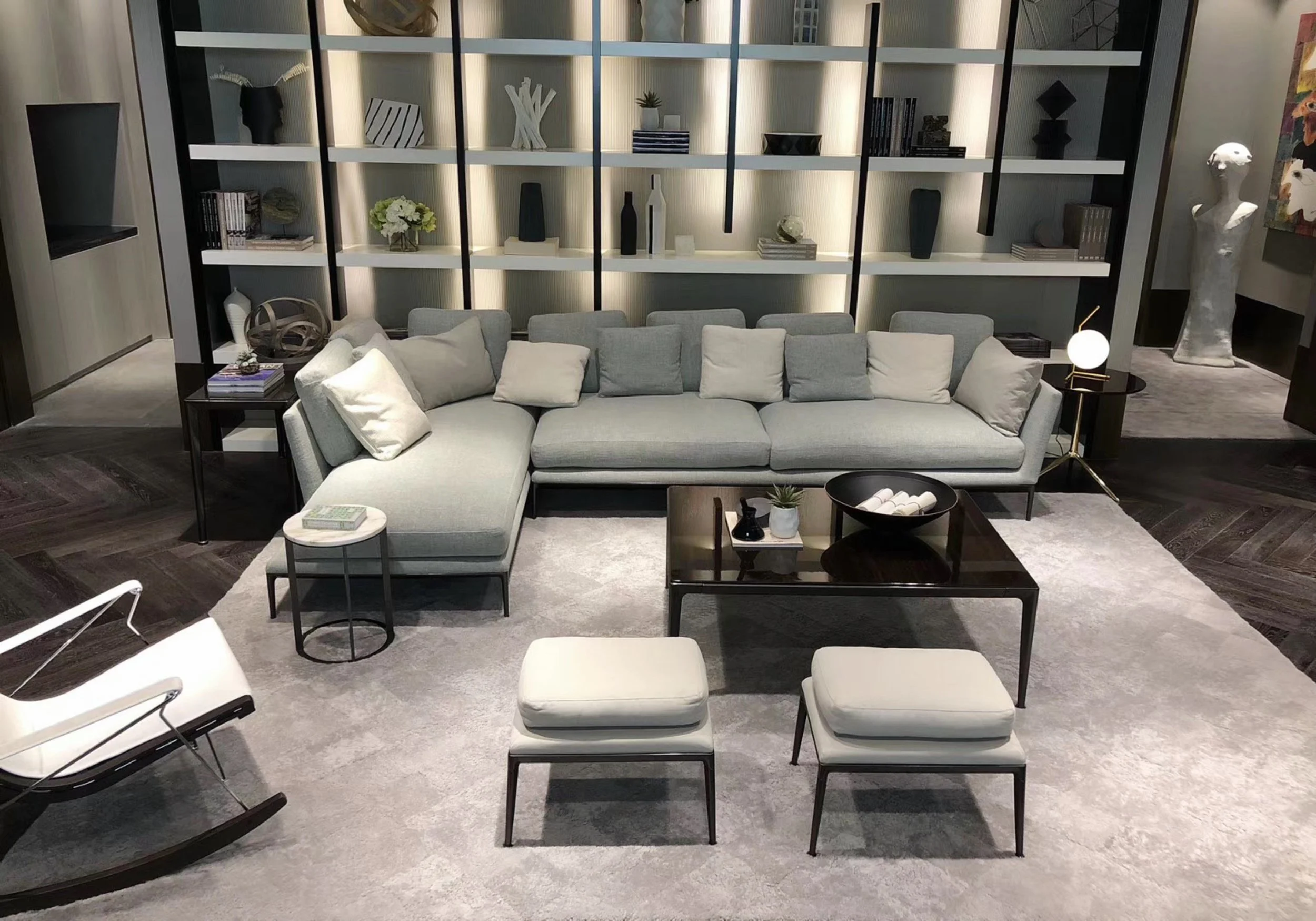 Modern gray sectional sofa and ottoman chairs in a stylish living room, with black shelving, decor, coffee table, rug, and warm lighting.