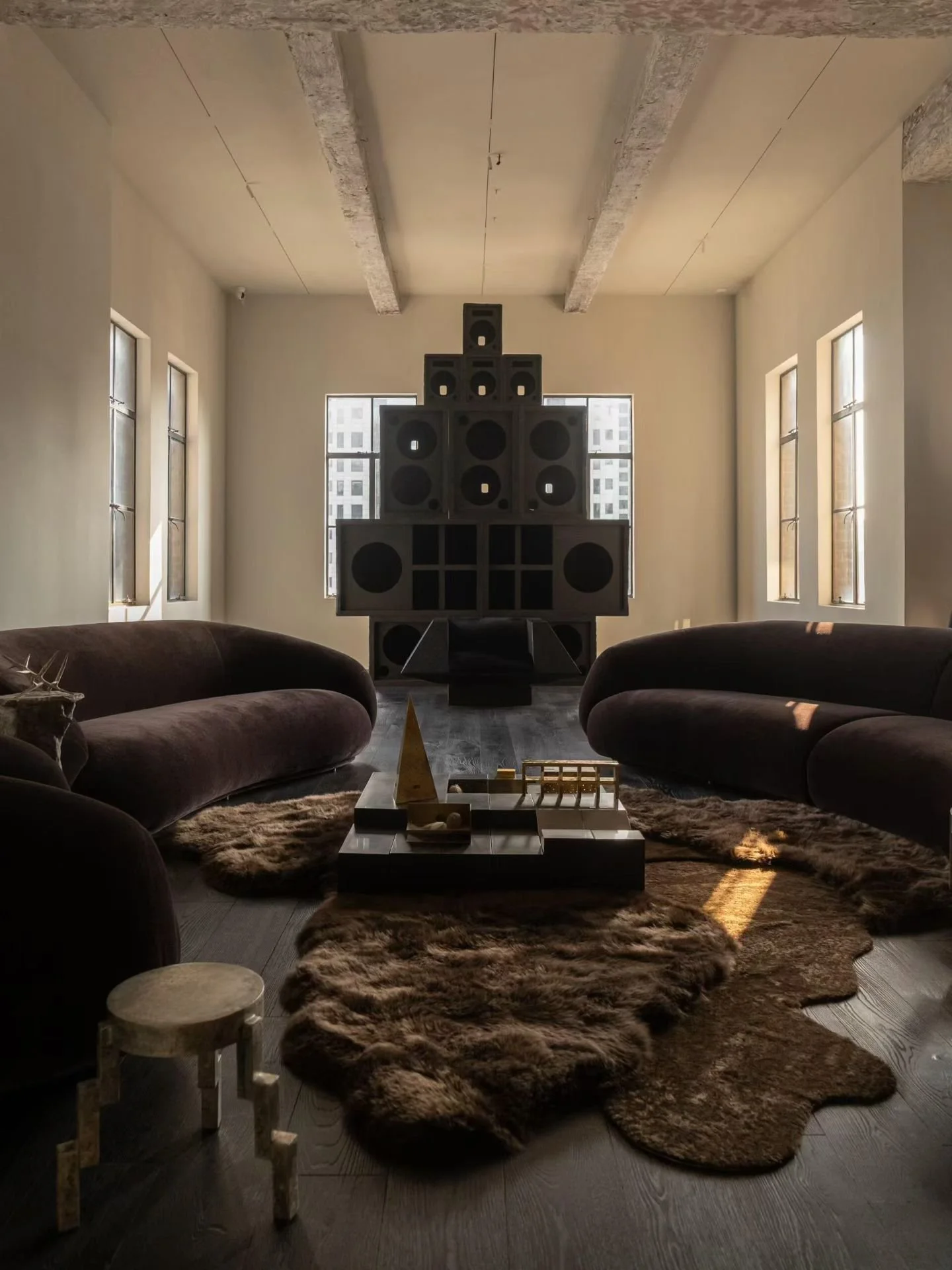 Moody modern living room with oversized vintage speakers, curved velvet sofas, chocolate fusion rug, coffee table, and warm sunlight through tall windows.
