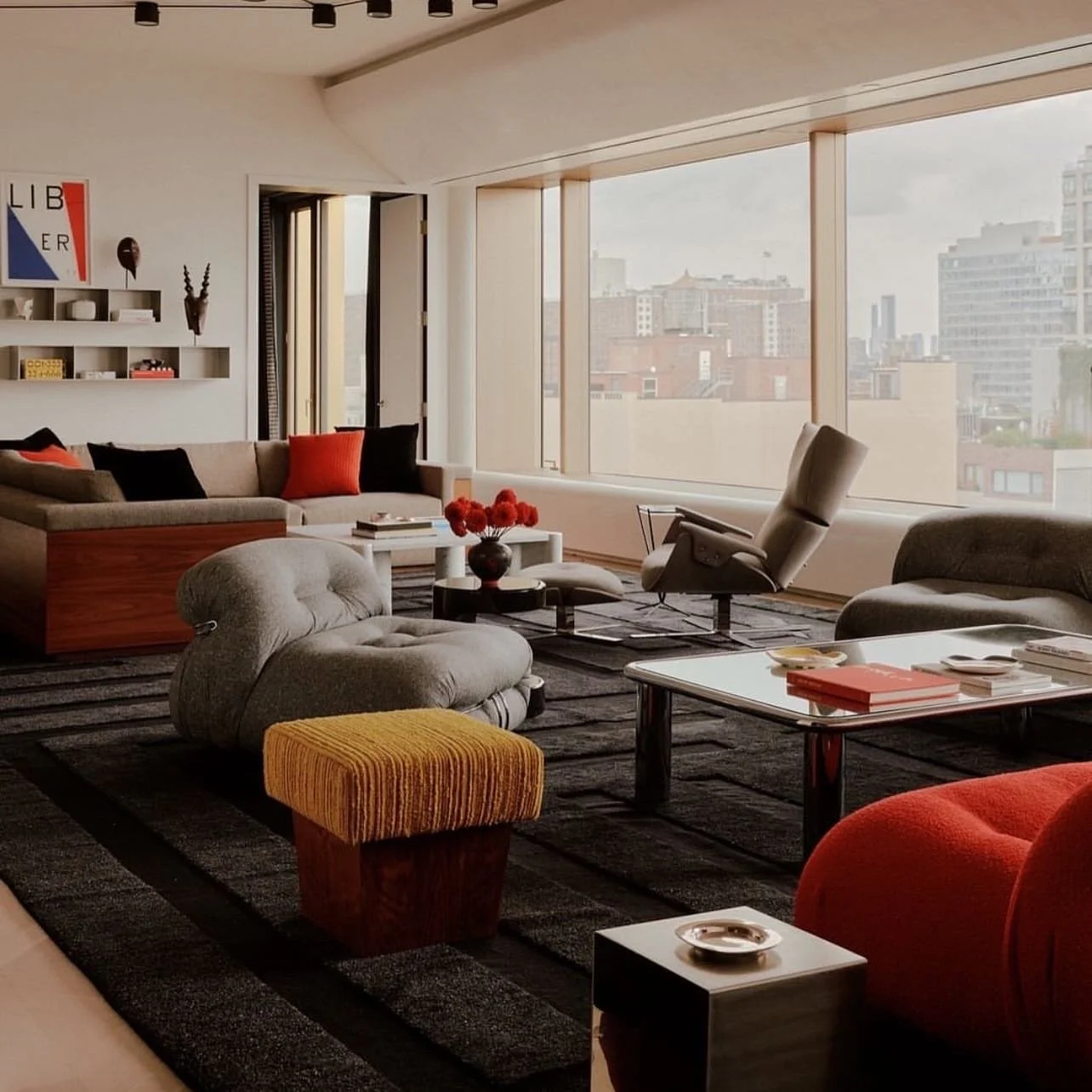 Modern living room with grey sofas, 3D design grey rug, red accents, lounge chair, coffee tables, and city views through large windows; stylish, inviting atmosphere.
