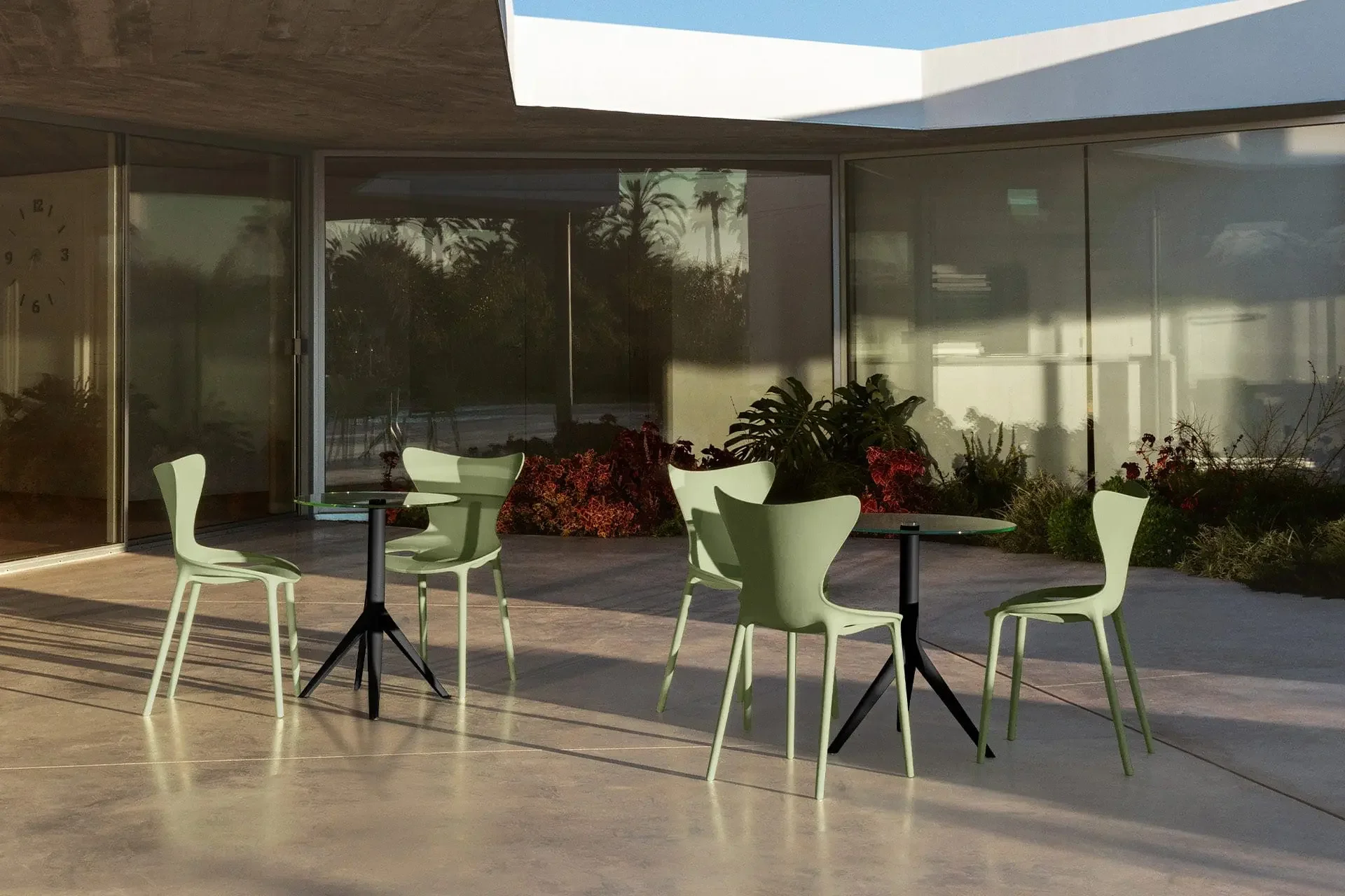 Sunlit modern patio with pale green chairs and black tables, glass walls, tropical plants, and long shadows creating a calm, inviting mood.