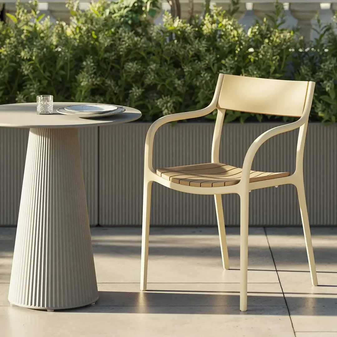 Modern outdoor patio scene with a cream armchair and ribbed pedestal table set with plates, surrounded by lush greenery in warm sunlight.