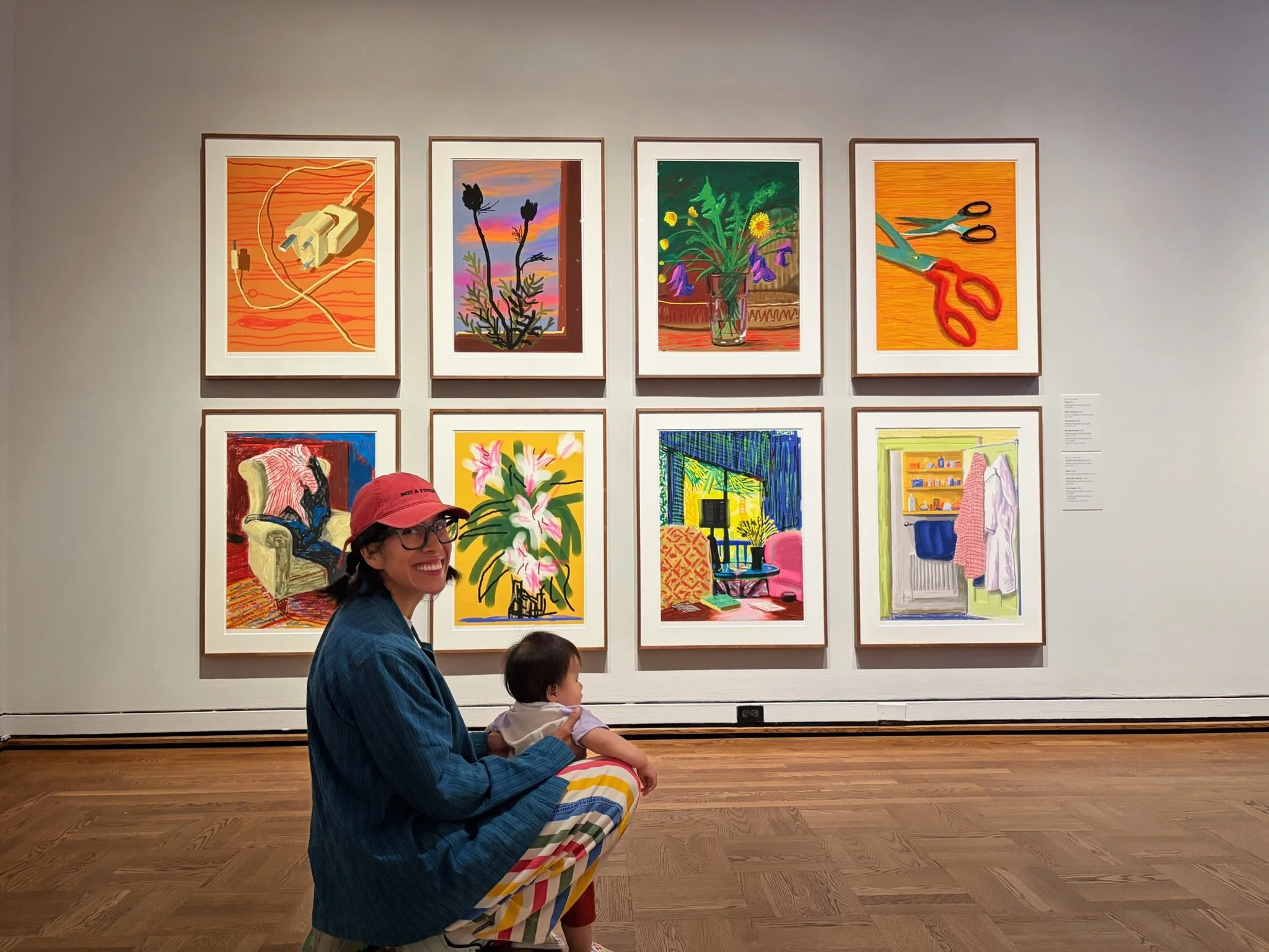 A woman wearing glasses and a red cap holding a young girl is smiling inside an art gallery. Behind them, a wall displays colorful contemporary paintings, including floral and indoor scenes.