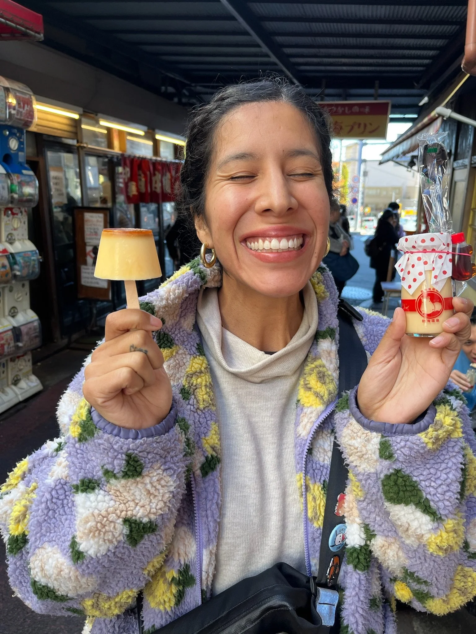 A woman with a broad smile holding a small dessert topped with caramel. She wears a colorful, fluffy jacket with floral patterns, gold hoop earrings, and a gray turtleneck sweater. The background appears to be a shopping or market area with vending machines and people.