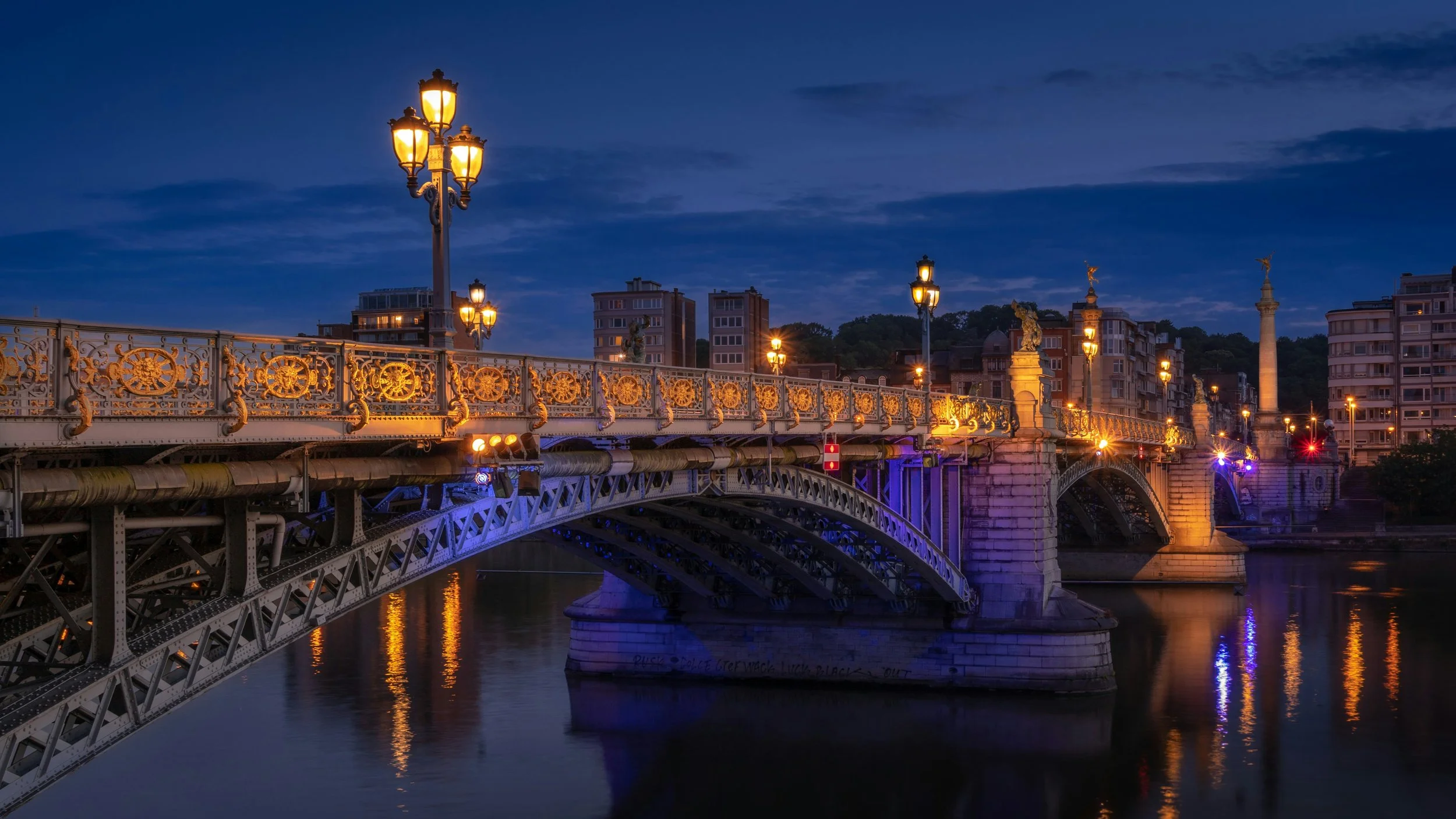 Night view of a decorative illuminated bridge over a river with city buildings and a hill in the background.