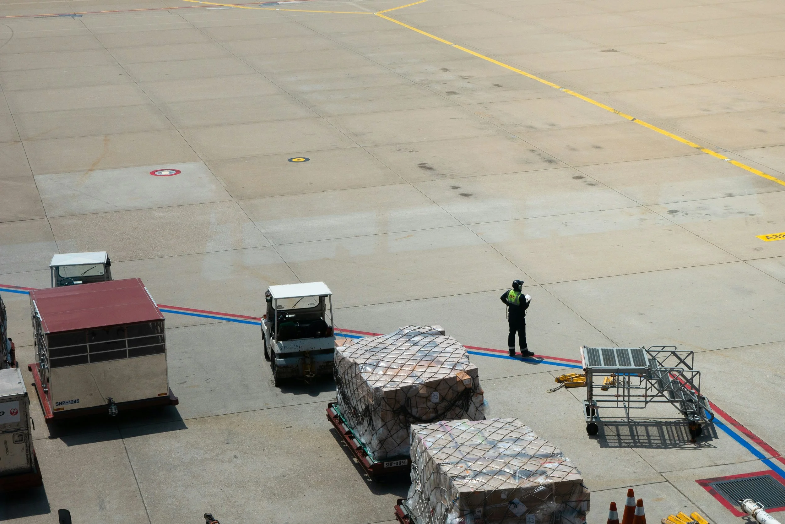Airport tarmac with ground crew, luggage carts, and cargo containers.