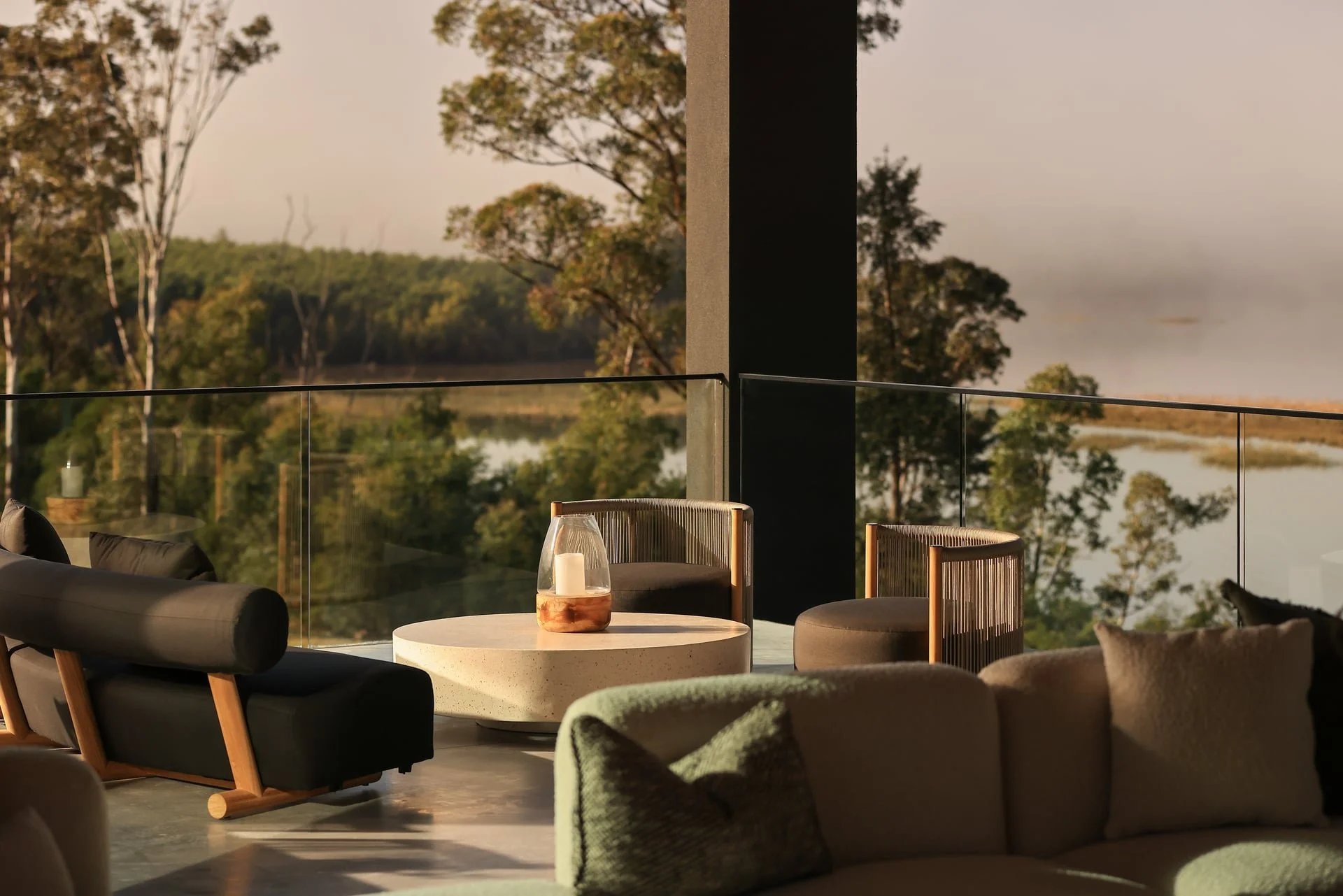 Indoor seating area with modern furniture and glass railing, overlooking a river and trees during sunset.