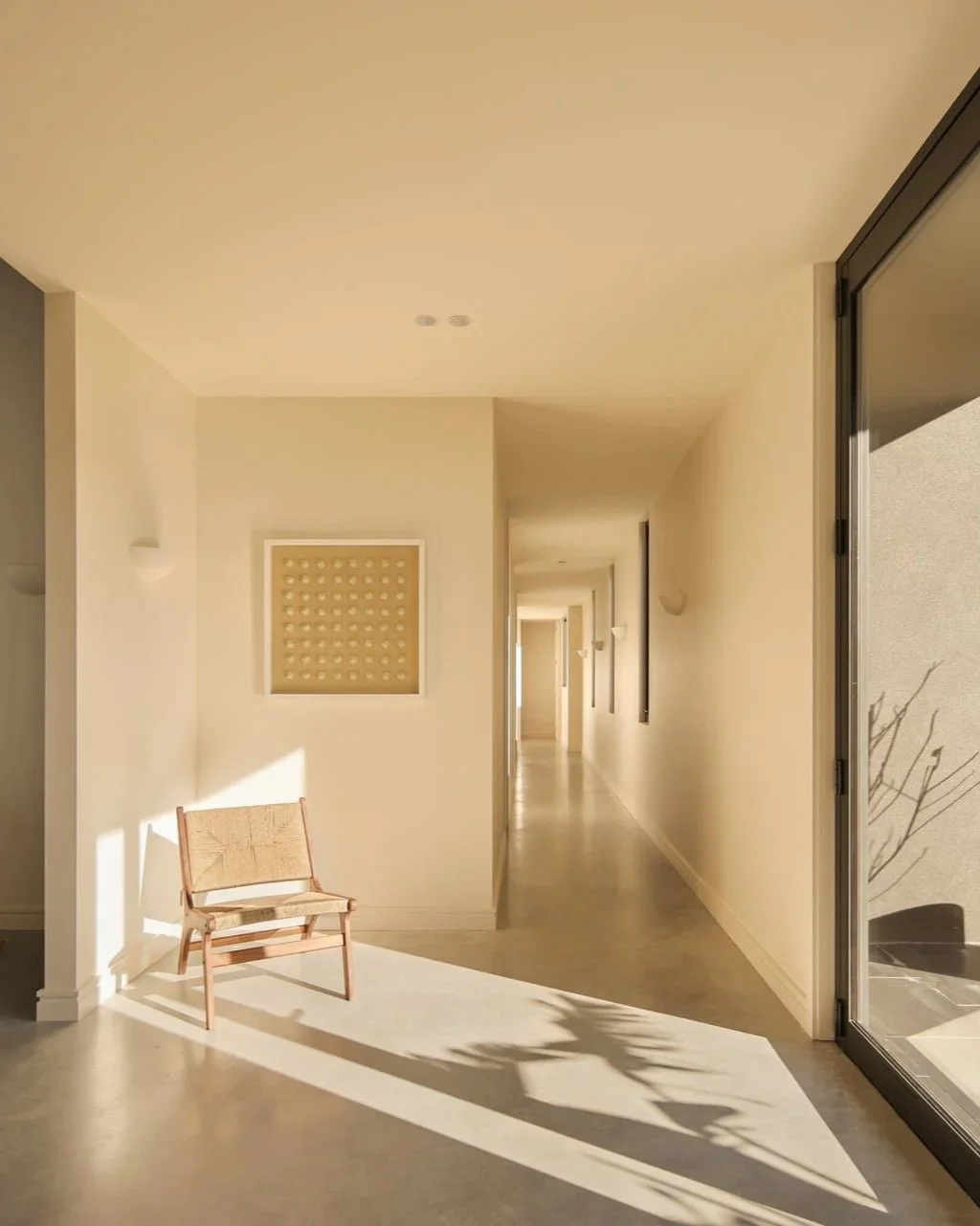 Sunlit hallway in a modern home with cream-colored walls, a single wooden chair, and large glass sliding doors opening to an outdoor patio.