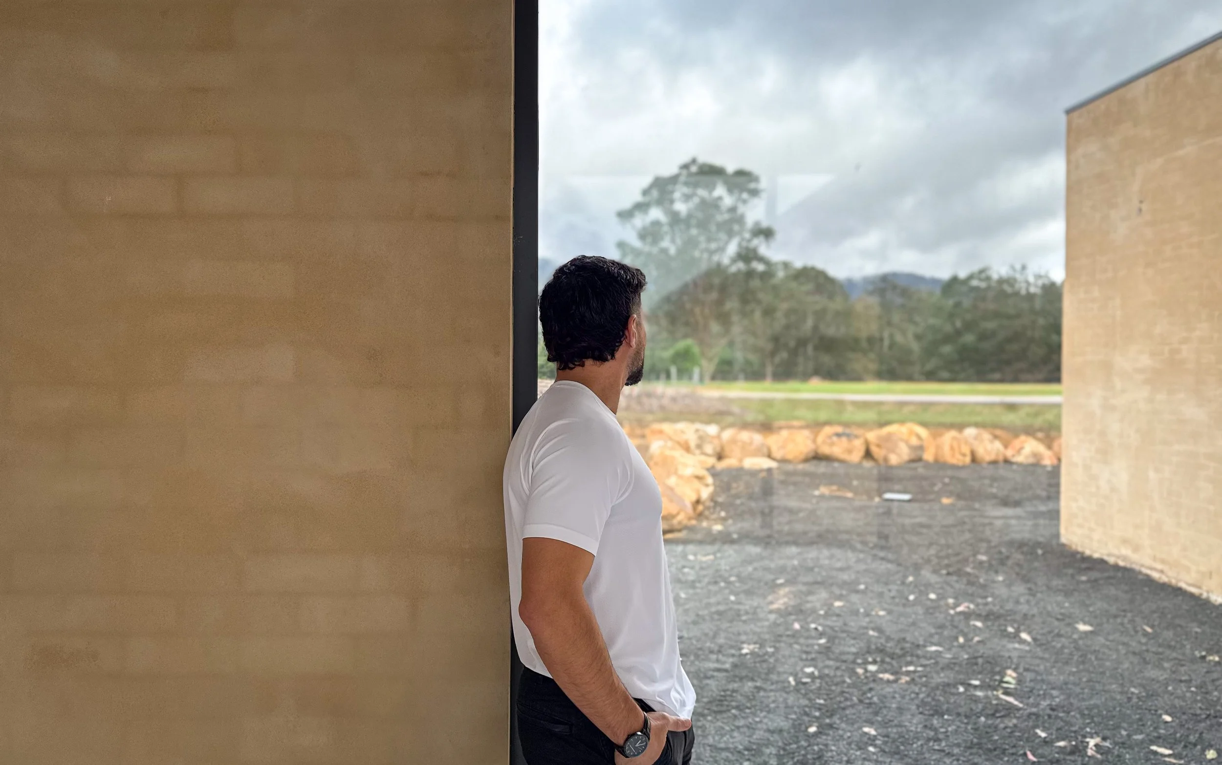 Alfie Arcuri, architect wearing a white t-shirt and black watch stands facing away and looking to the side. He is positioned near a beige wall with natural scenery visible through an open space, including trees, cloudy sky, and rocks on the ground.