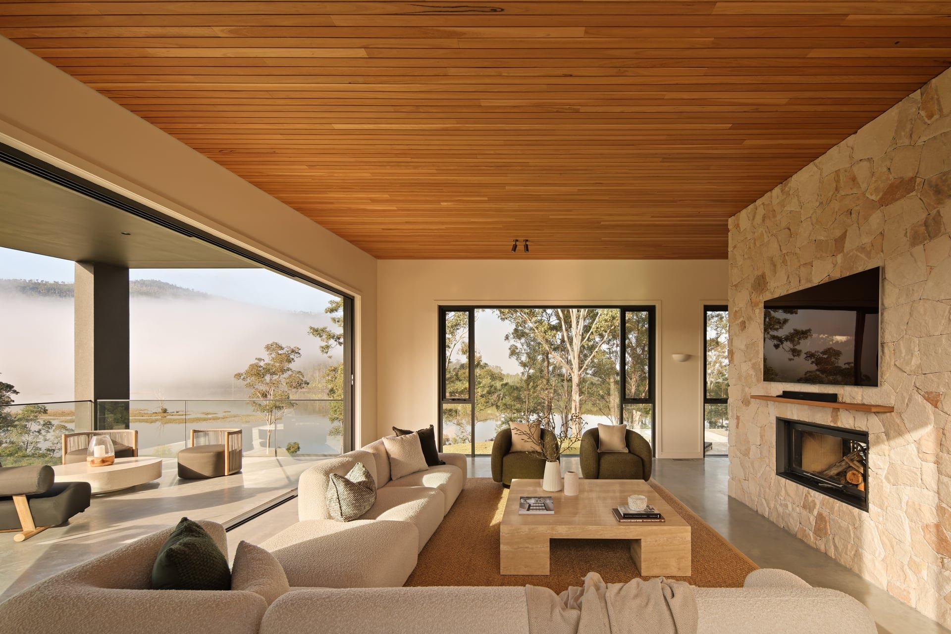 Modern living room with large windows, wood-paneled ceiling, stone fireplace, and scenic outdoor view of trees and water.
