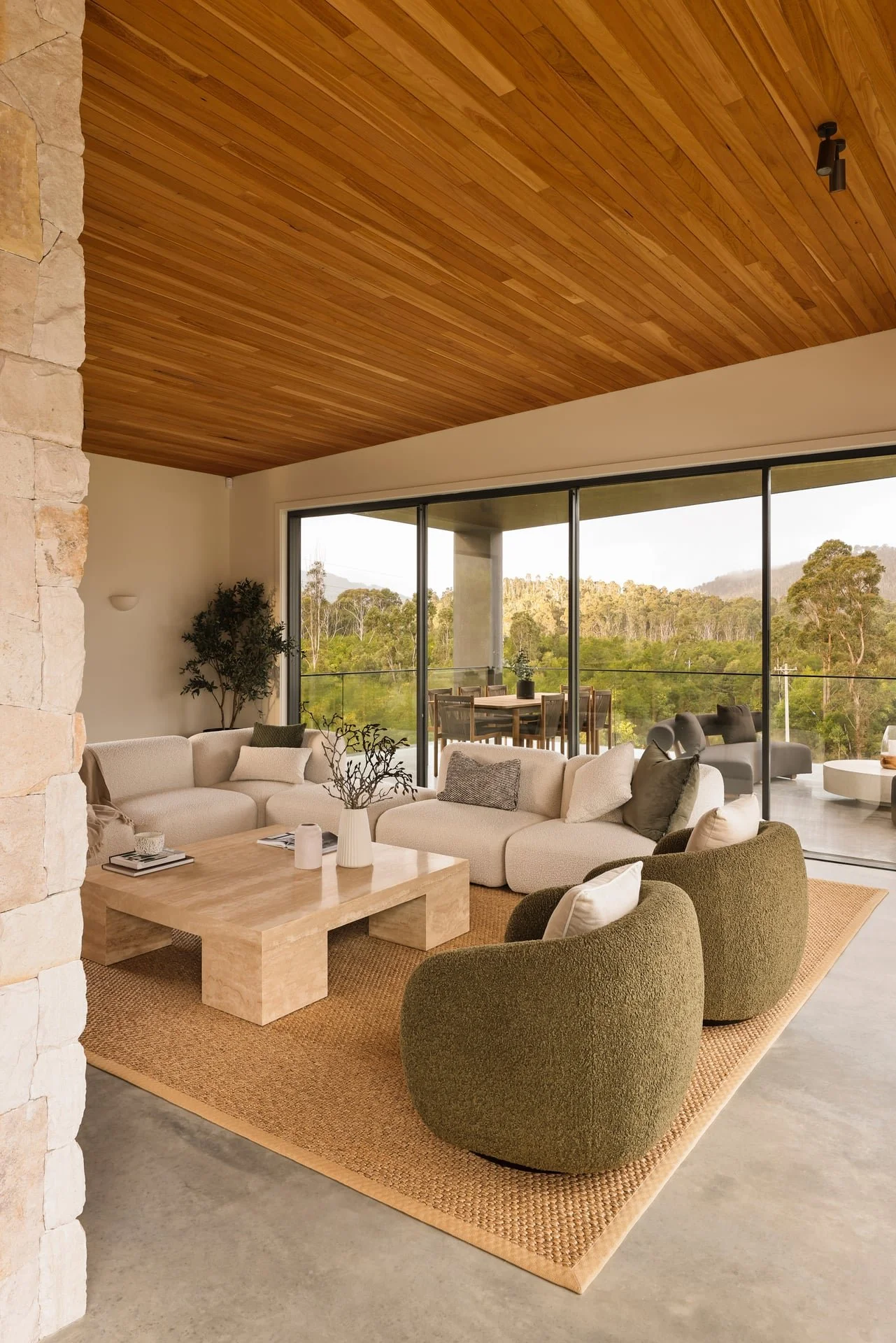 Living room with a beige sectional sofa, green armchairs, a wooden coffee table with vases, a beige rug, large glass sliding doors opening to a balcony with outdoor seating, overlooking a lush green landscape with trees and hills, and a wooden ceiling.