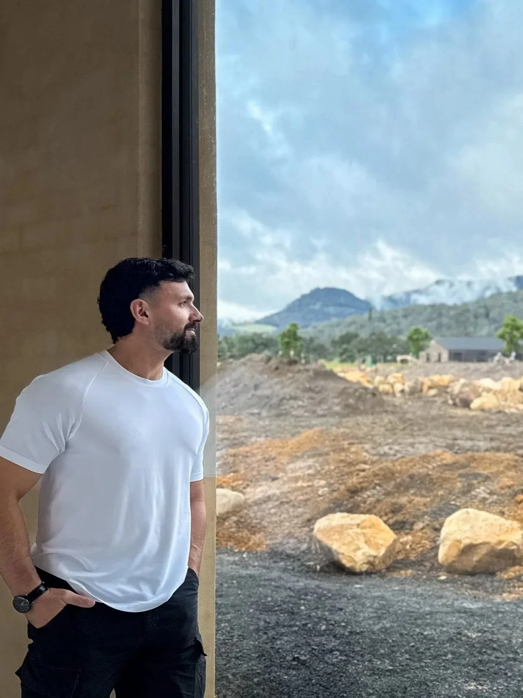 Alfie Arcuri the architect, wearing a white T-shirt and black pants, stands indoors with his hands in his pockets, looking out a large window at a mountainous landscape with clouds and rocks.
