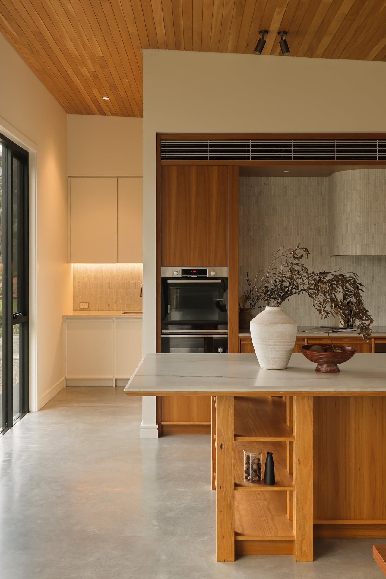 Modern kitchen with wooden cabinetry, a built-in oven, white countertops, a large white vase with dried branches, and a small wooden shelf with decorative items. Wooden ceiling with track lighting, beige walls, and sliding glass doors on the left.