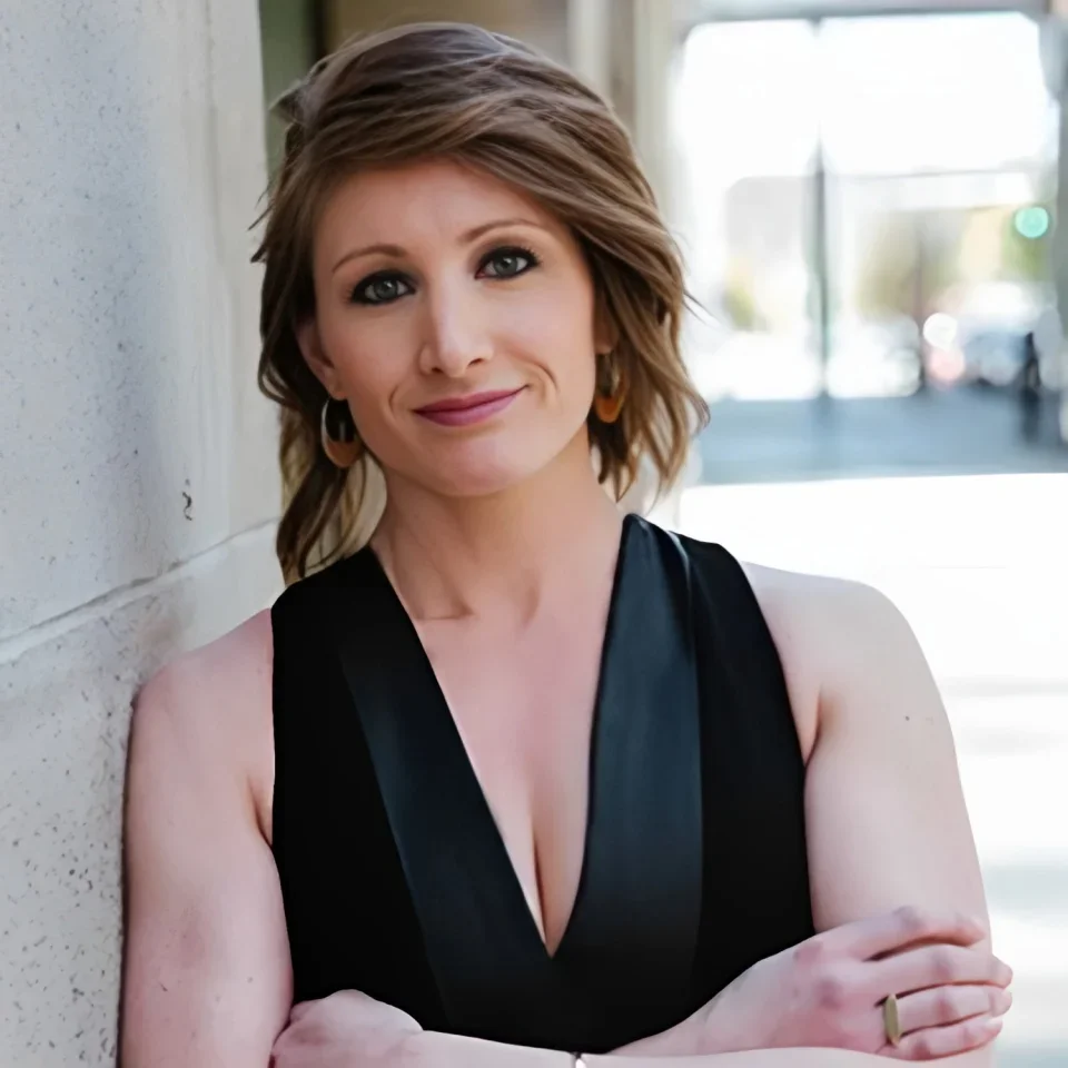 A woman with short, wavy brown hair, wearing a black sleeveless top, gold hoop earrings, and a ring on her right hand, leaning against a light-colored wall outdoors.