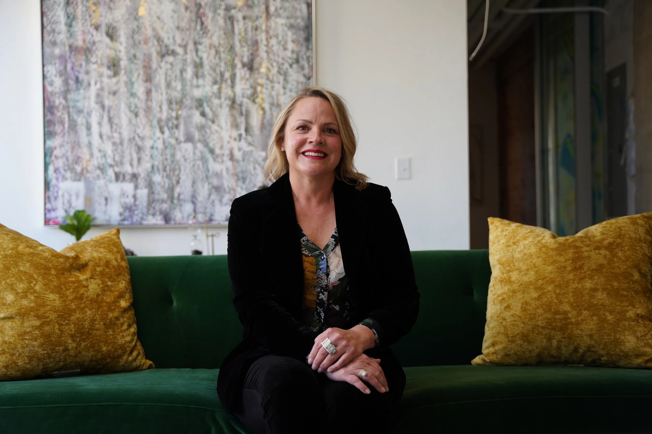 A woman with blonde hair smiling and sitting on a green velvet couch, surrounded by yellow velvet pillows, in a modern living space with abstract artwork on the wall behind her.