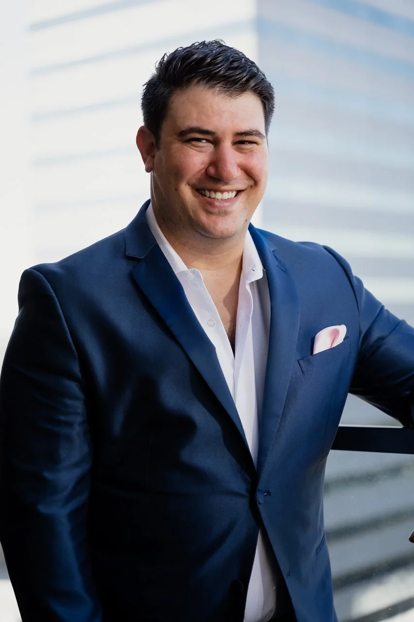 Man in a navy blue suit and white shirt smiling at the camera, standing outdoors with a modern building in the background.